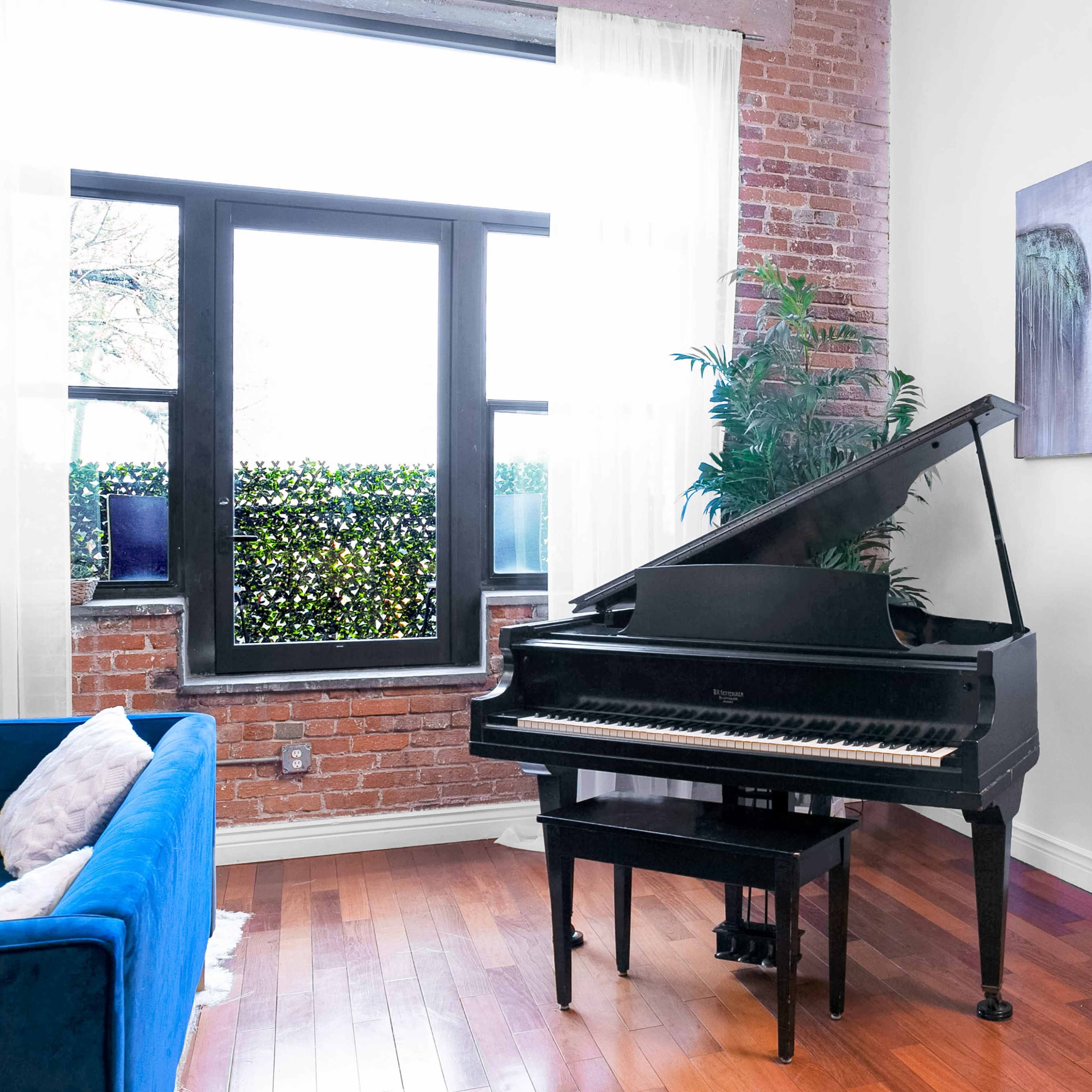 A black grand piano is positioned near a window in a room with brick walls and wooden floors, accompanied by two abstract paintings and a small plant.