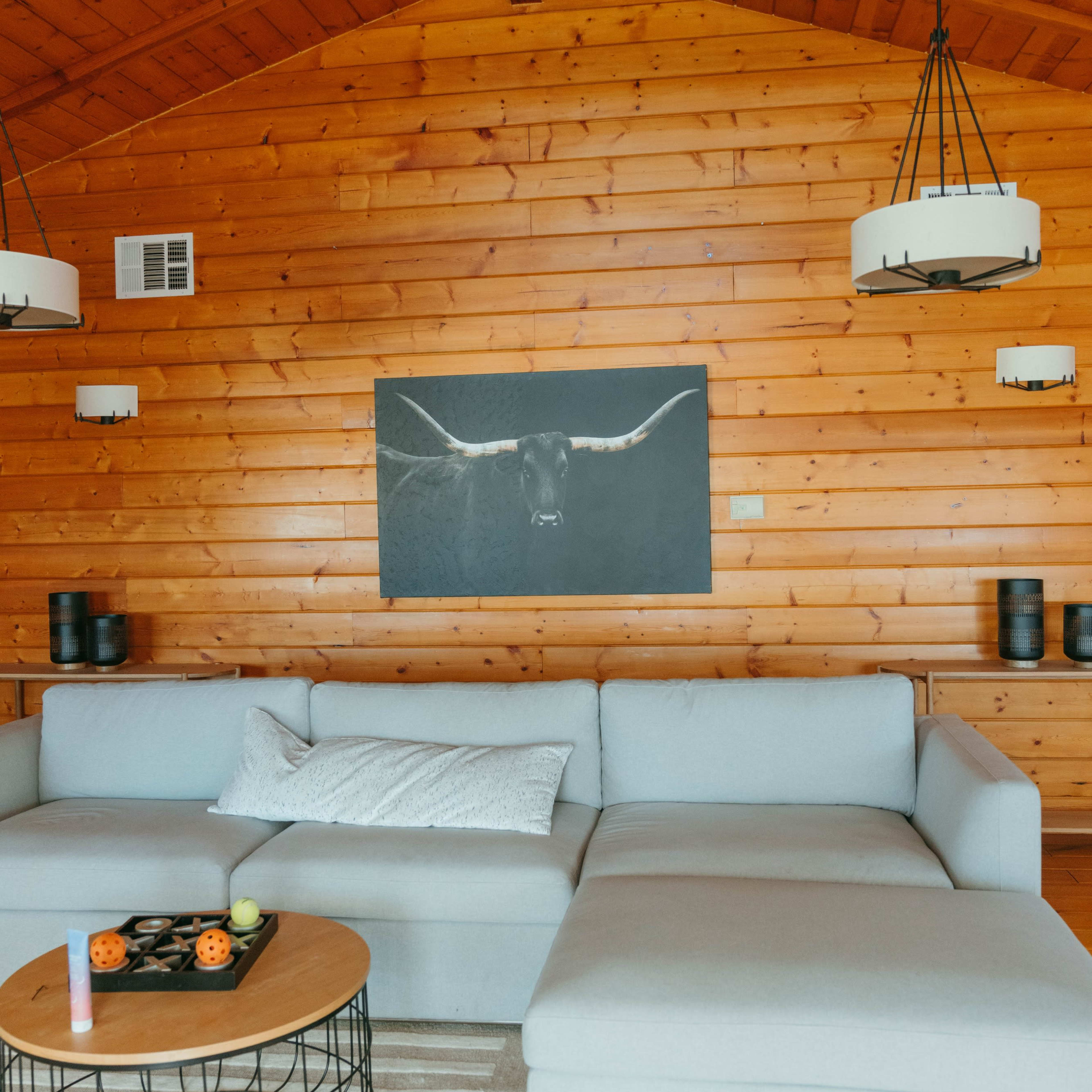 A living room with a wooden paneled wall, a large light-colored sectional sofa, a round coffee table, and a painting of a bull's head above the sofa.