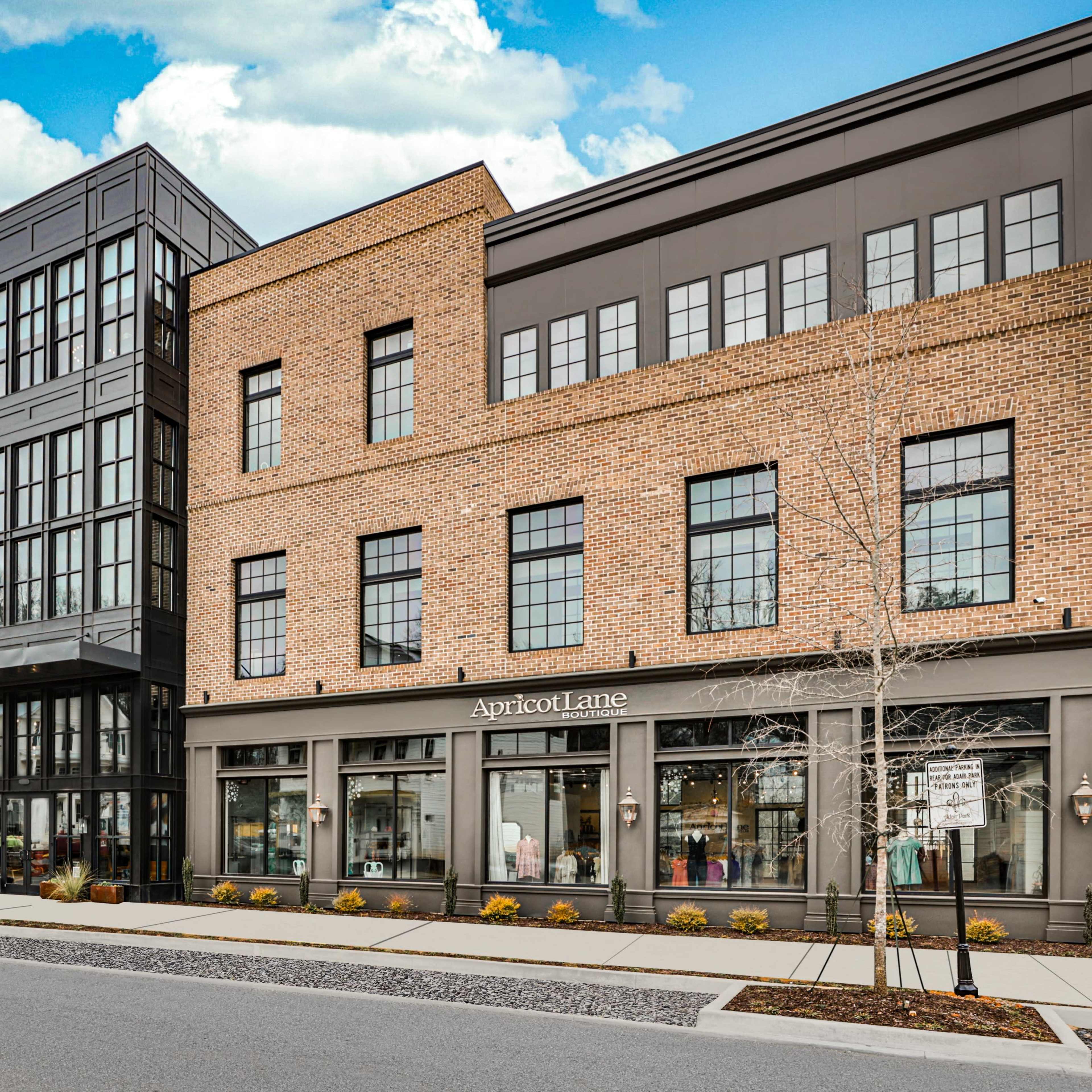 A modern multi-story brick building with large glass windows and retail spaces on the ground floor, located along a street lined with planters.