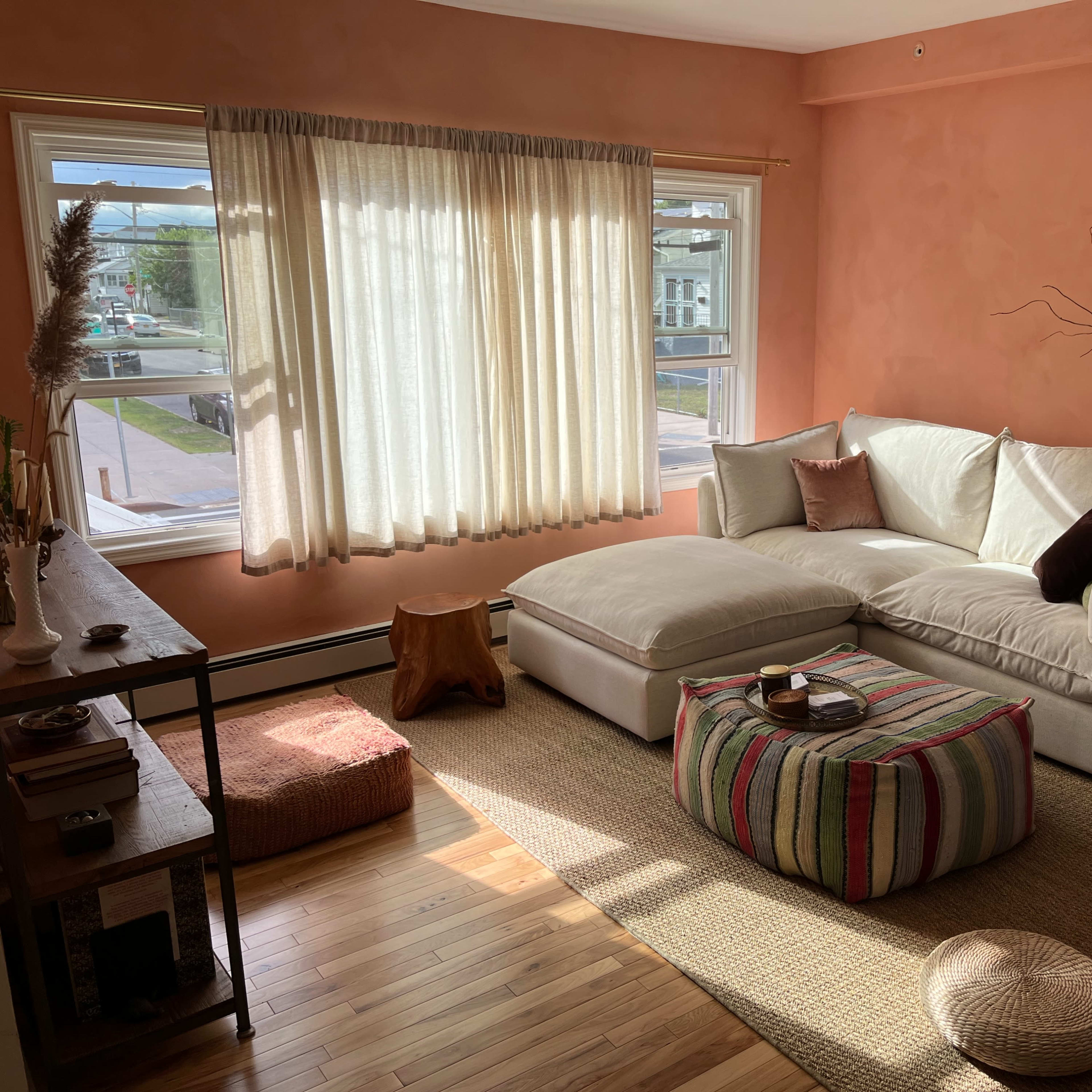 A cozy living room with a large white sectional sofa, a wooden coffee table, woven decor, and a large window with sheer curtains letting in natural light.