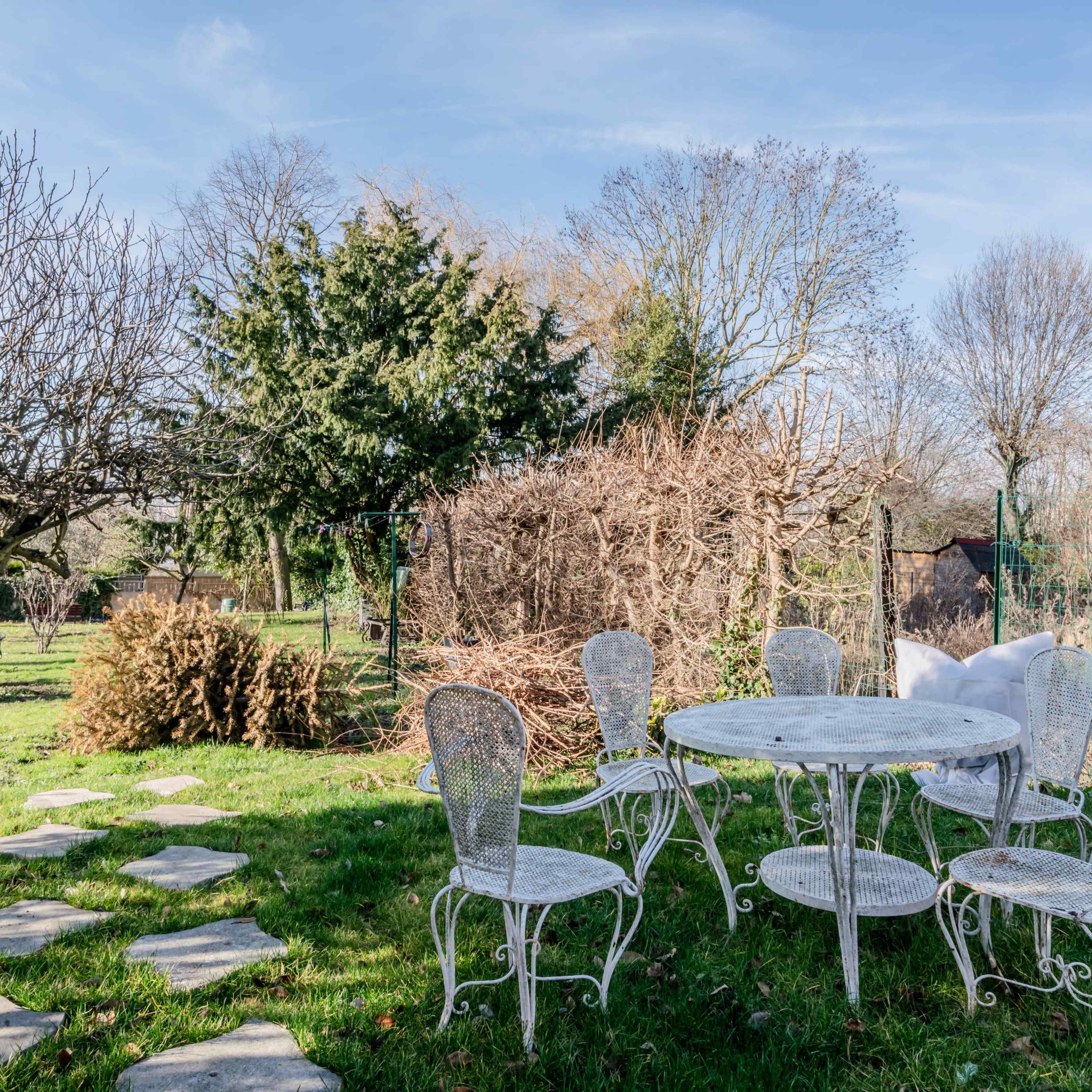 A garden with a circular table surrounded by six chairs, featuring a stone pathway and various trees in the background.