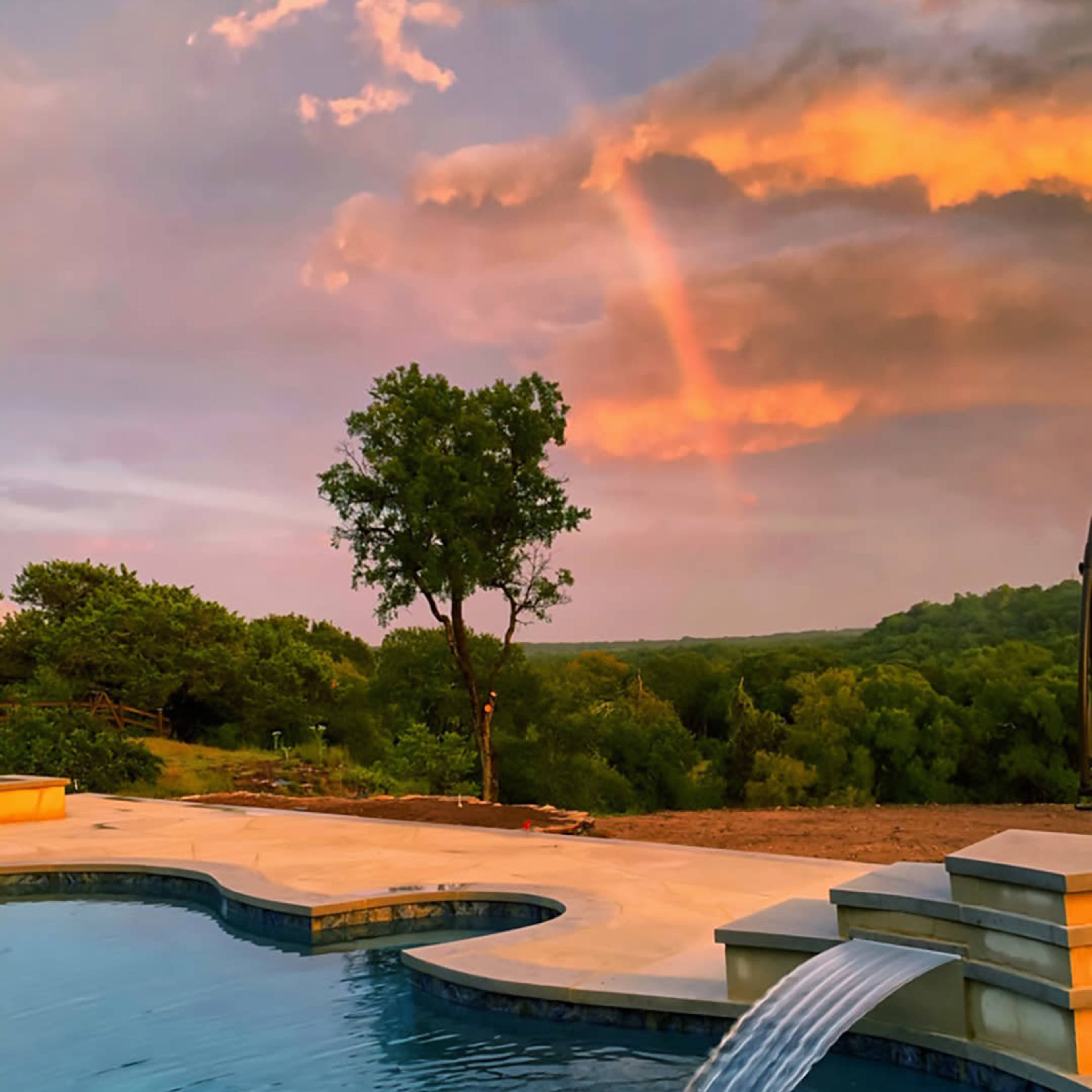 A swimming pool features a cascading water wall, set against a backdrop of green trees and a rainbow amidst colorful clouds.