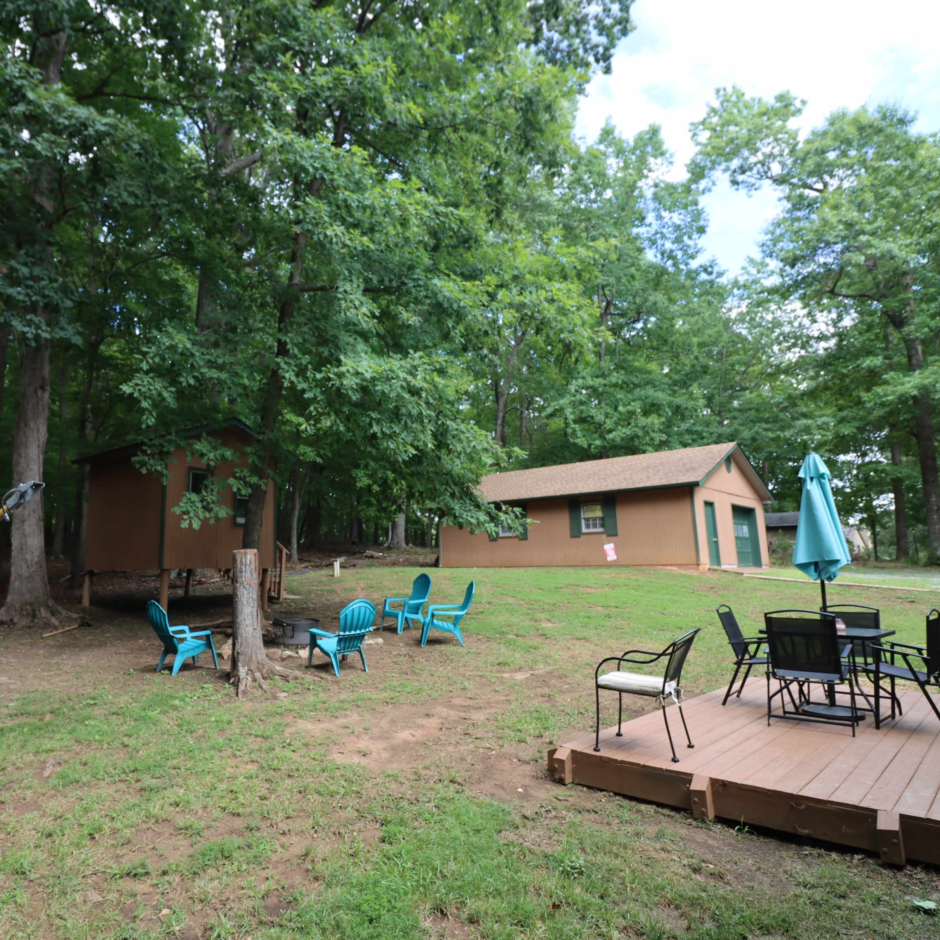 A wooded area featuring a brown building, a small structure, a satellite dish, and outdoor seating with a patio table and chairs.
