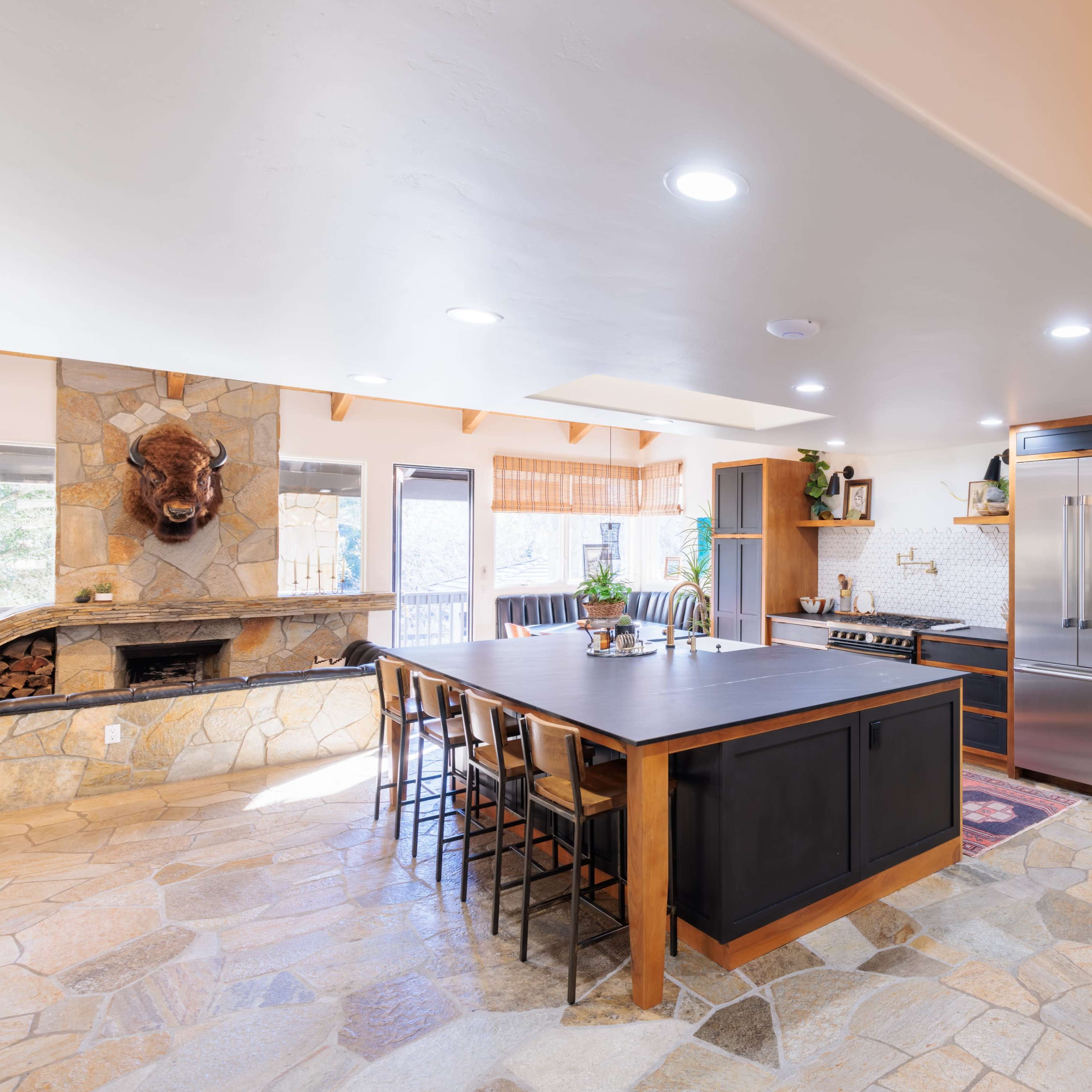 A modern kitchen with a large central island, stainless steel appliances, a stone fireplace, and a combination of wooden and stone flooring.