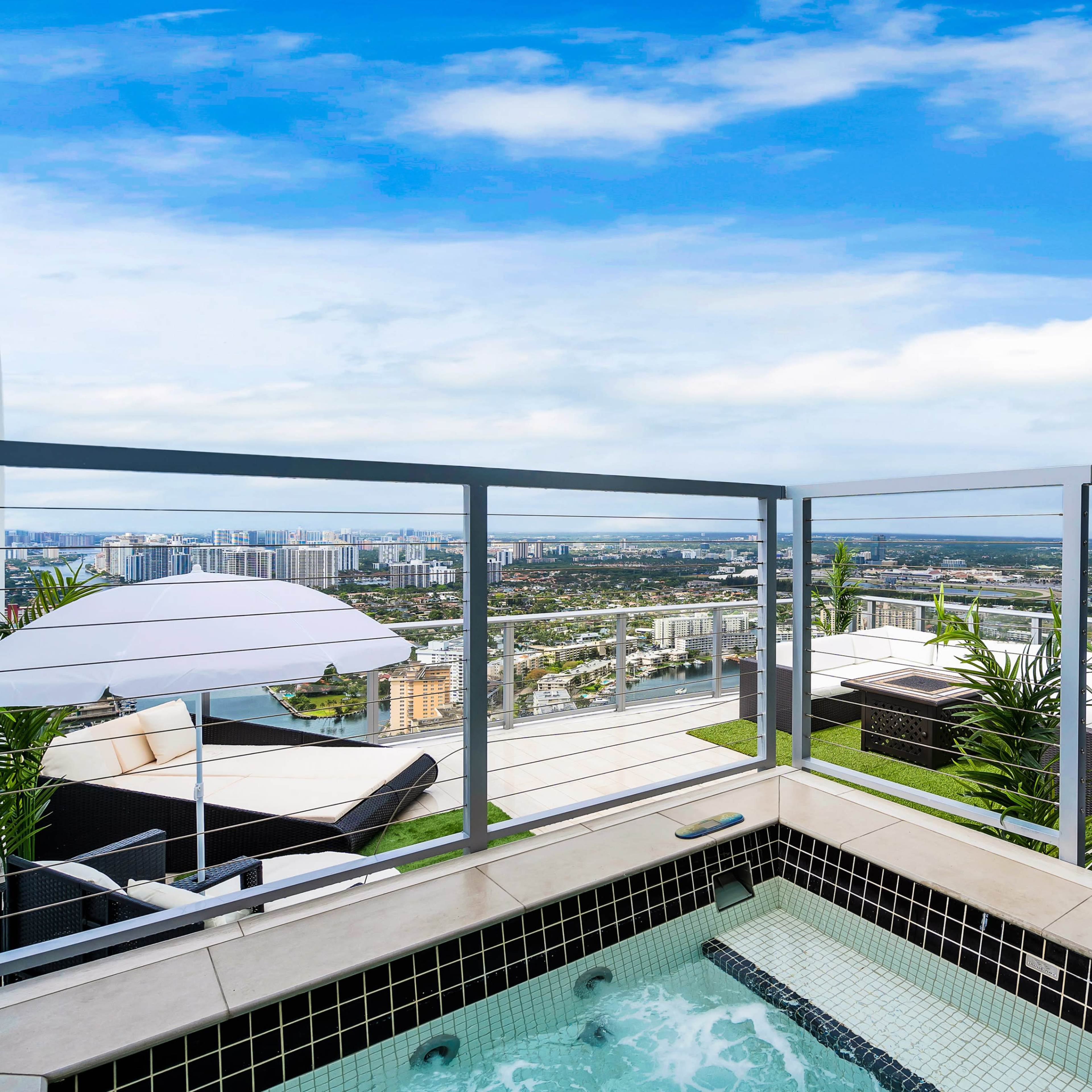 A rooftop terrace featuring a hot tub, lounge chairs, and a panoramic view of a cityscape under a cloudy sky.
