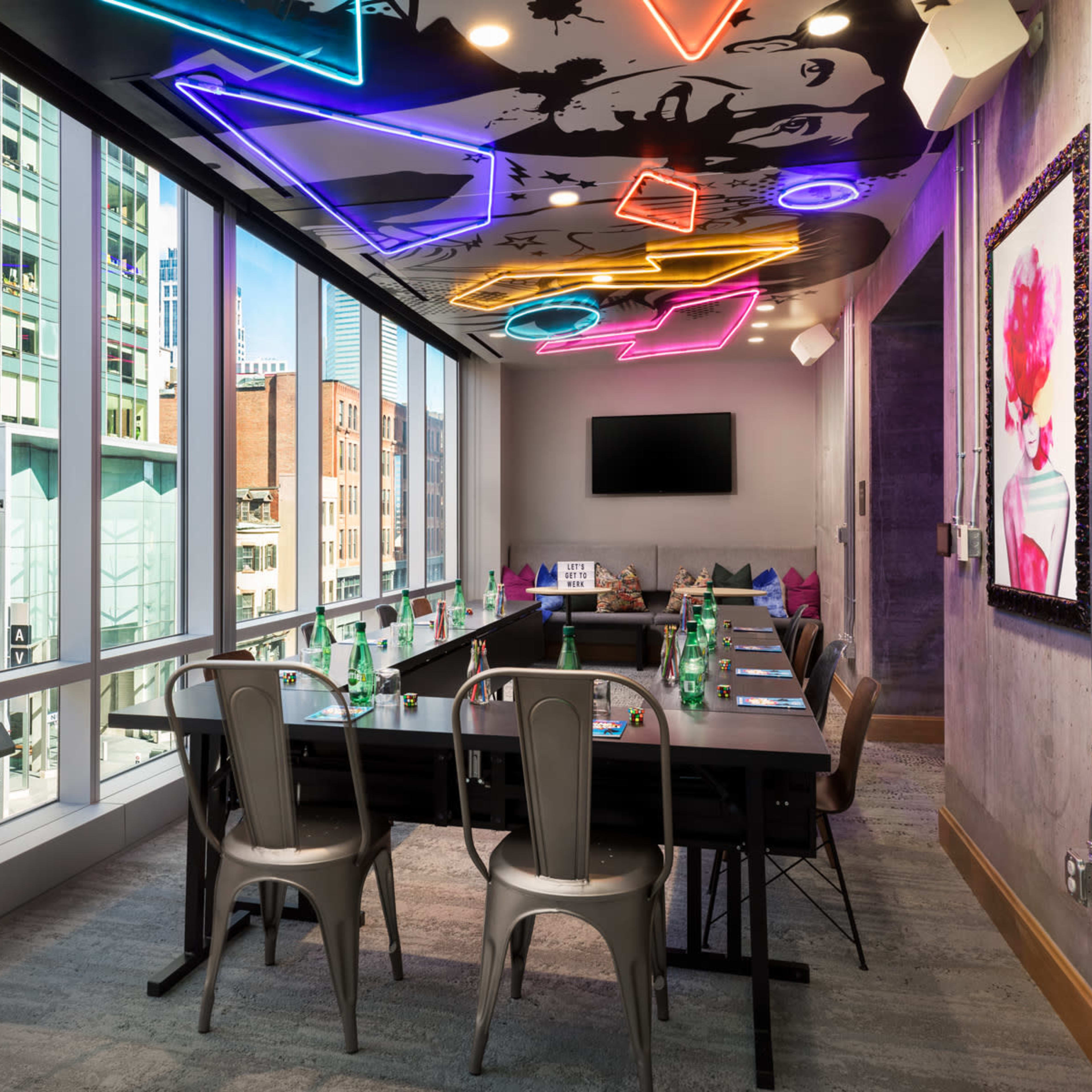 A modern conference room features large windows with city views, a long dark table, and colorful neon lighting on the ceiling.