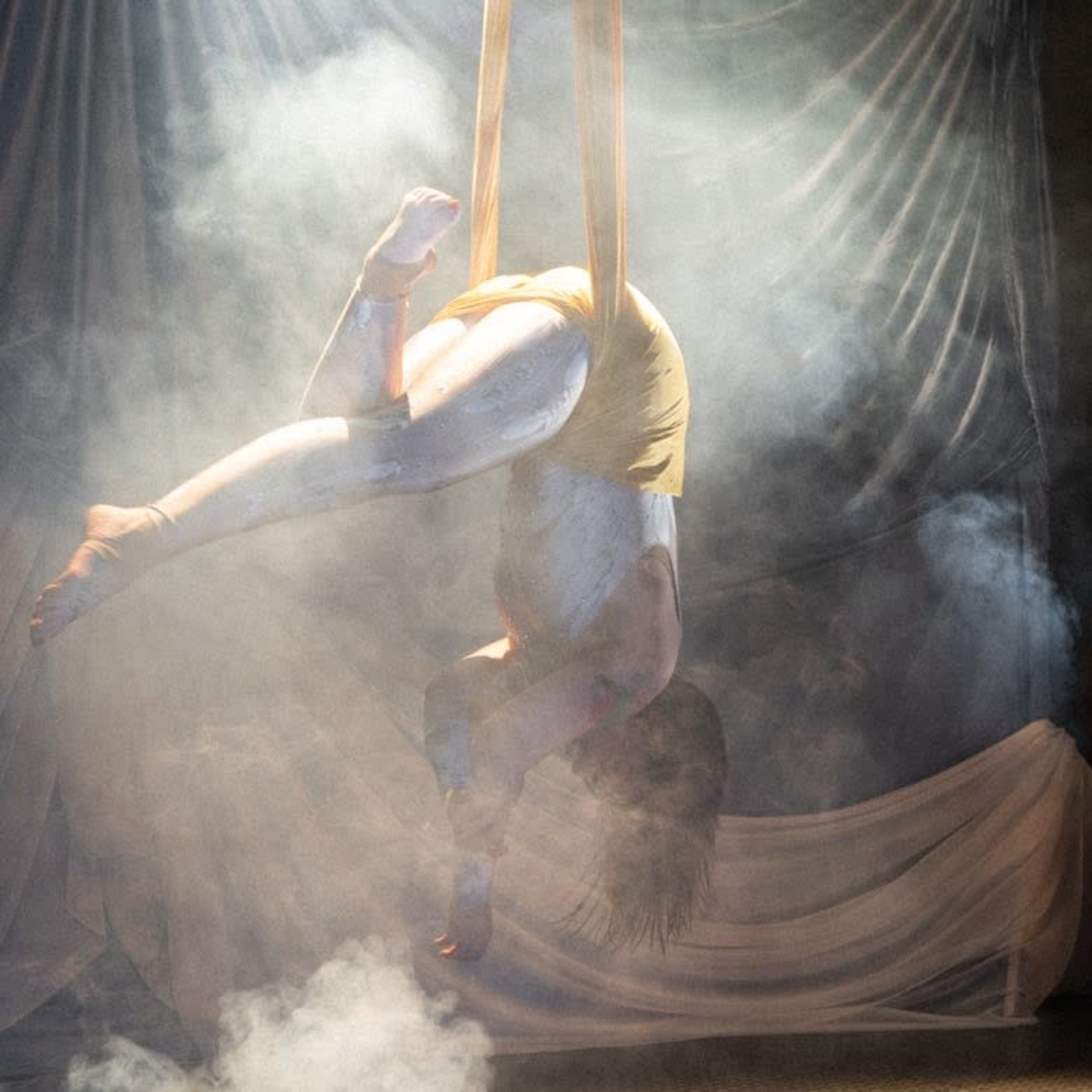 A performer is suspended in the air using fabric while a woman captures the scene with a camera, all amidst a backdrop of smoke and draped fabric.