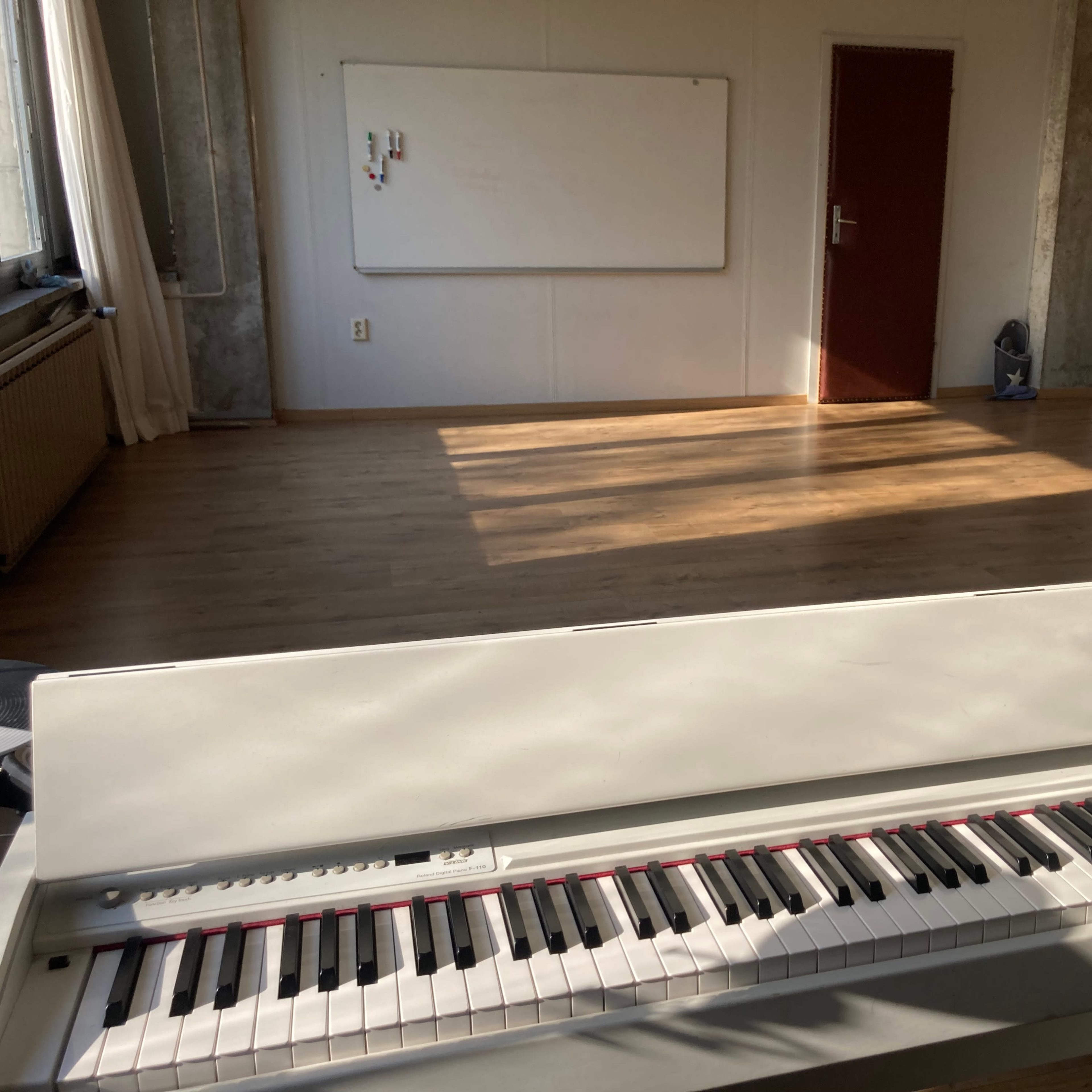 A white digital piano is positioned in the foreground of a sunlit room with wooden flooring and a whiteboard on the wall.