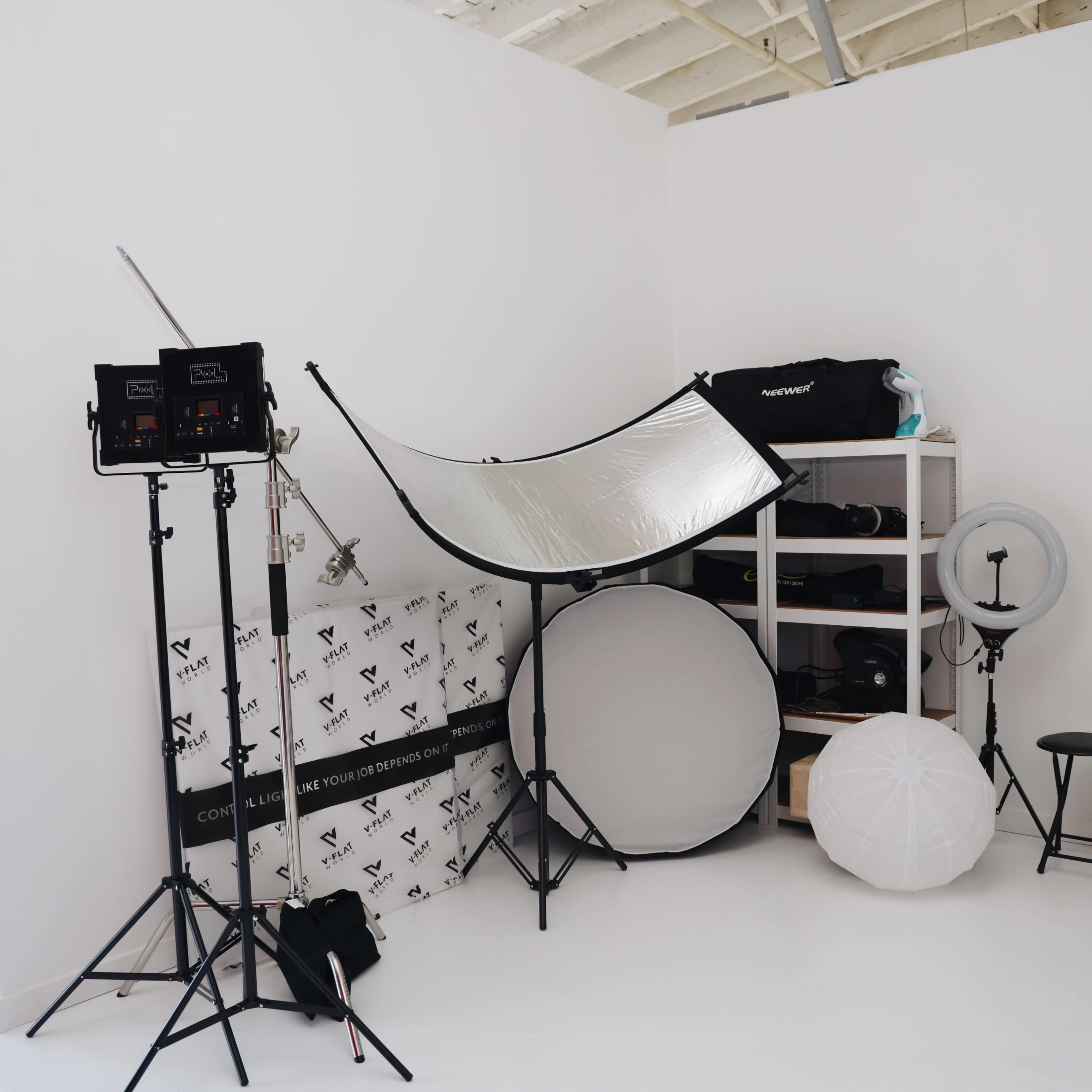 A photography studio corner with lighting equipment, reflectors, a shelving unit, and various backdrops against a white wall.