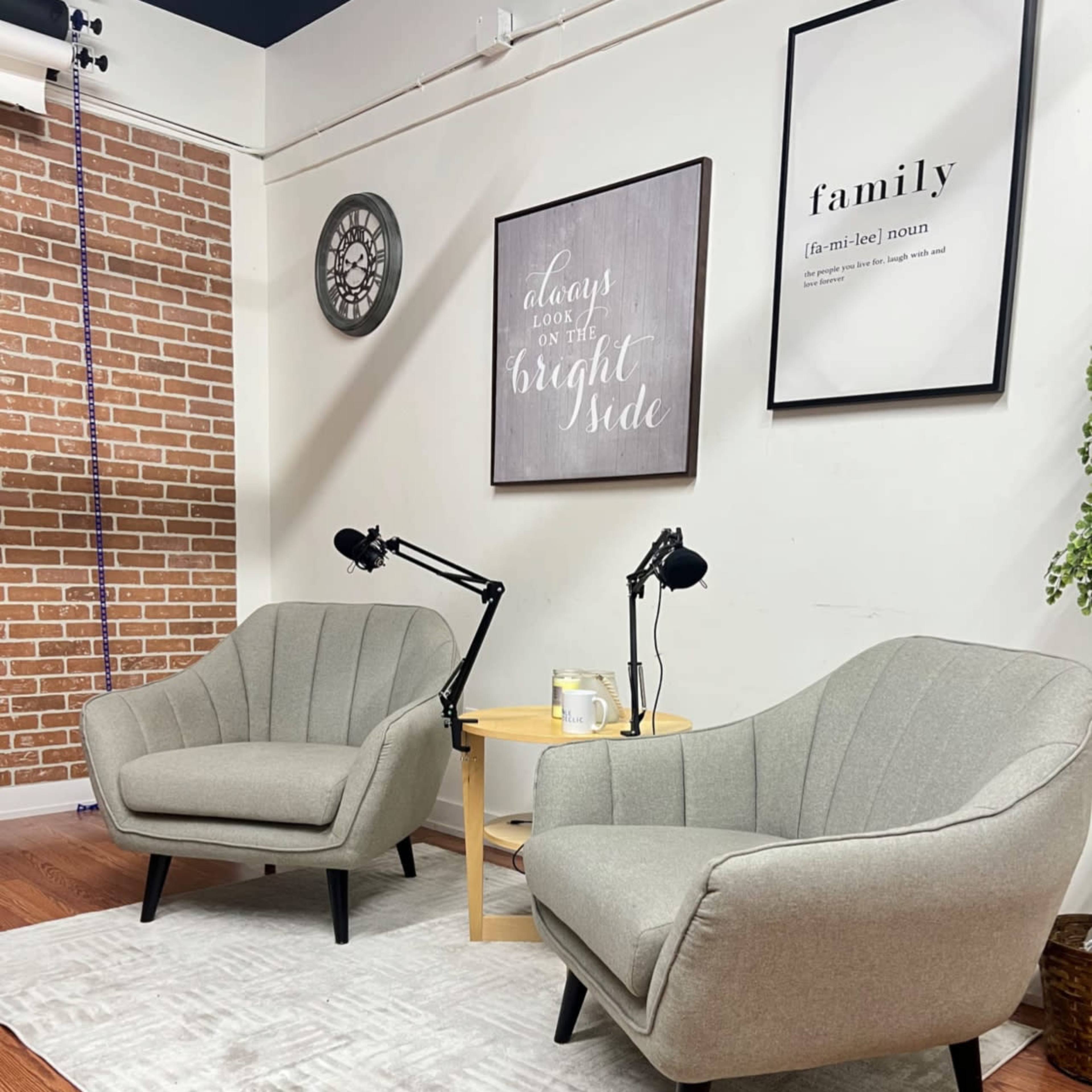 Two gray chairs facing each other on a light-colored rug, with a small round table and microphones positioned nearby in a softly decorated room.