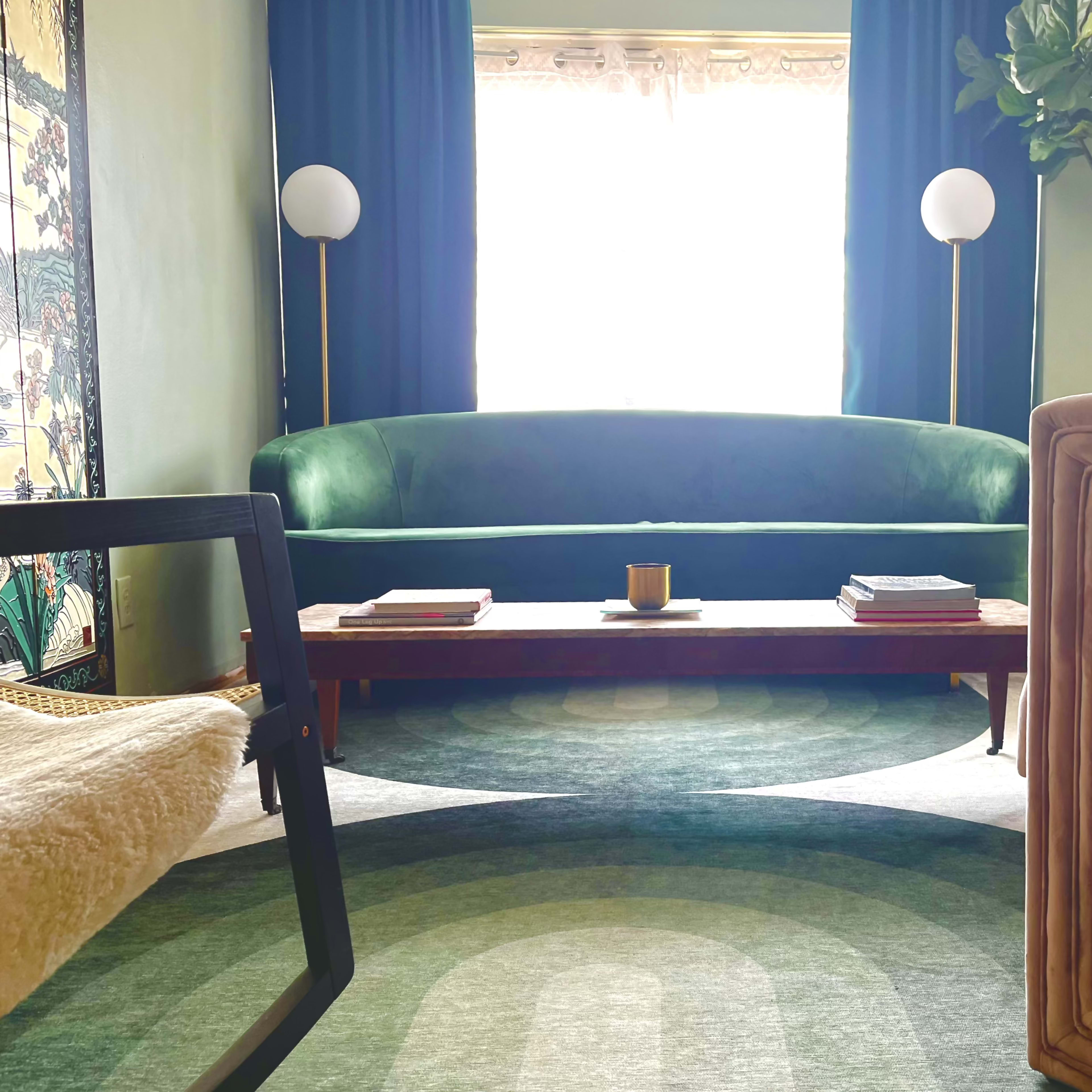 A green sofa and a beige armchair are positioned around a wooden coffee table on a patterned rug in a brightly lit living room.