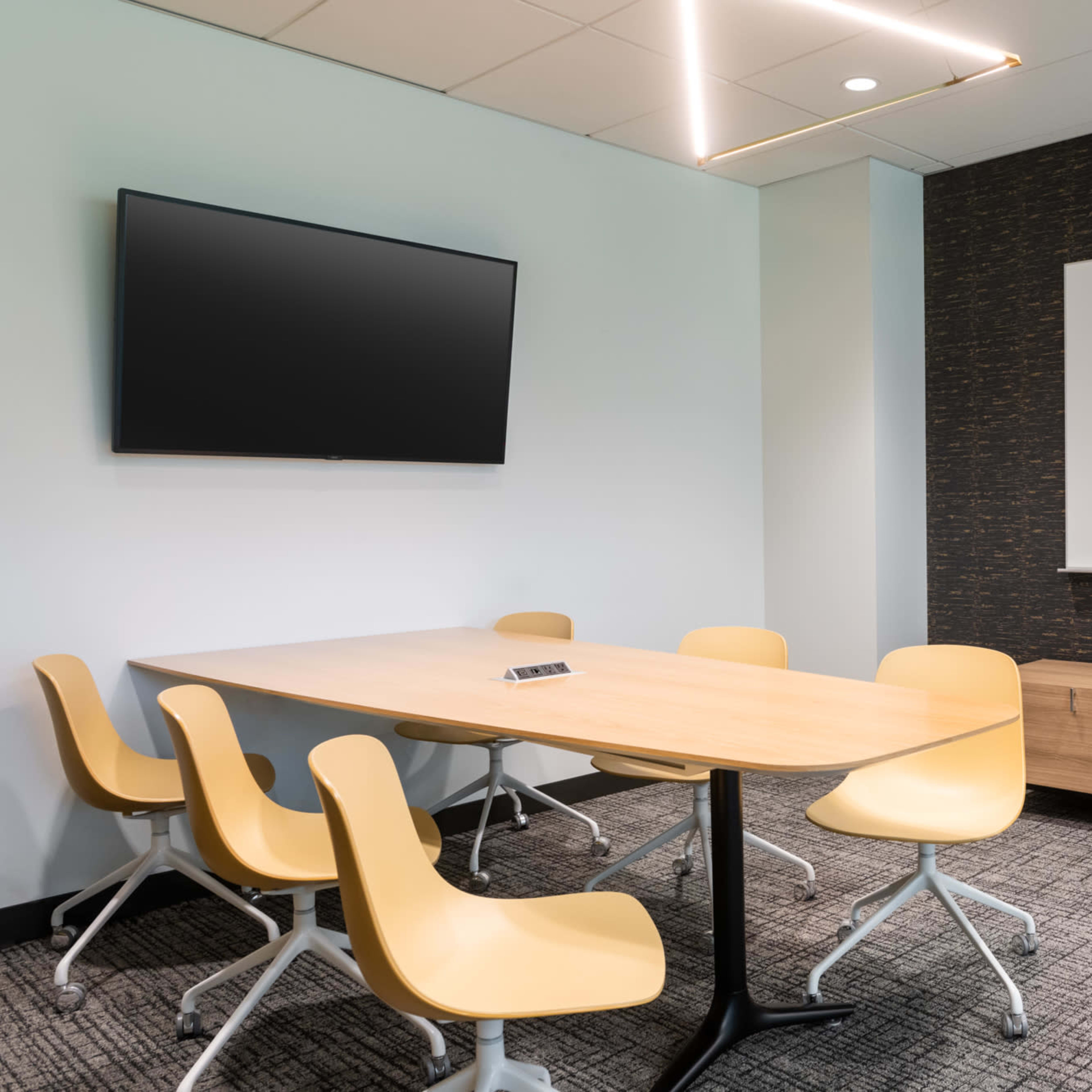 A modern meeting room features a wooden table surrounded by yellow chairs, a wall-mounted television, and a whiteboard on the dark-shaded wall.