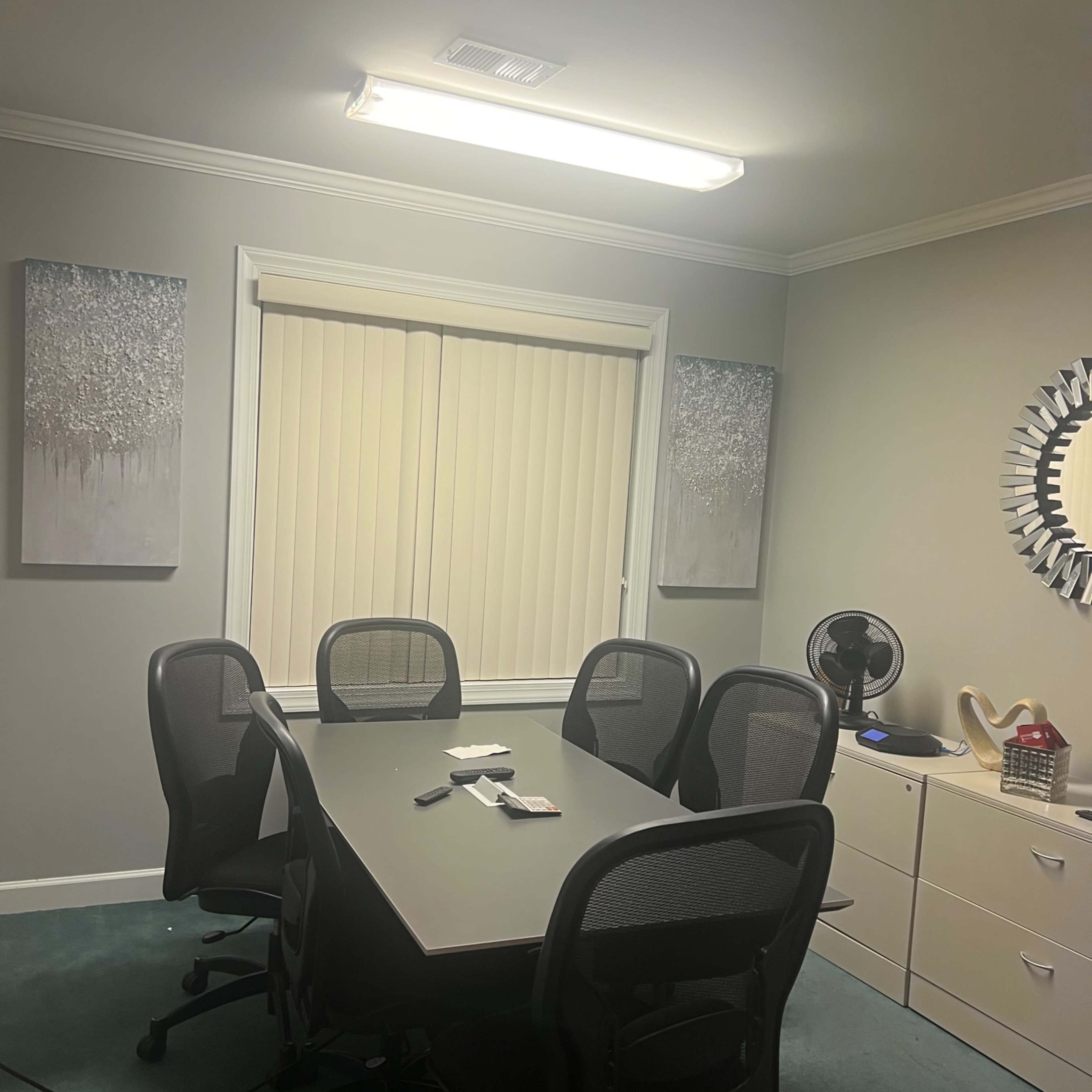 A conference room features a rectangular table surrounded by six chairs, with two paintings on the walls and a window covered by vertical blinds.