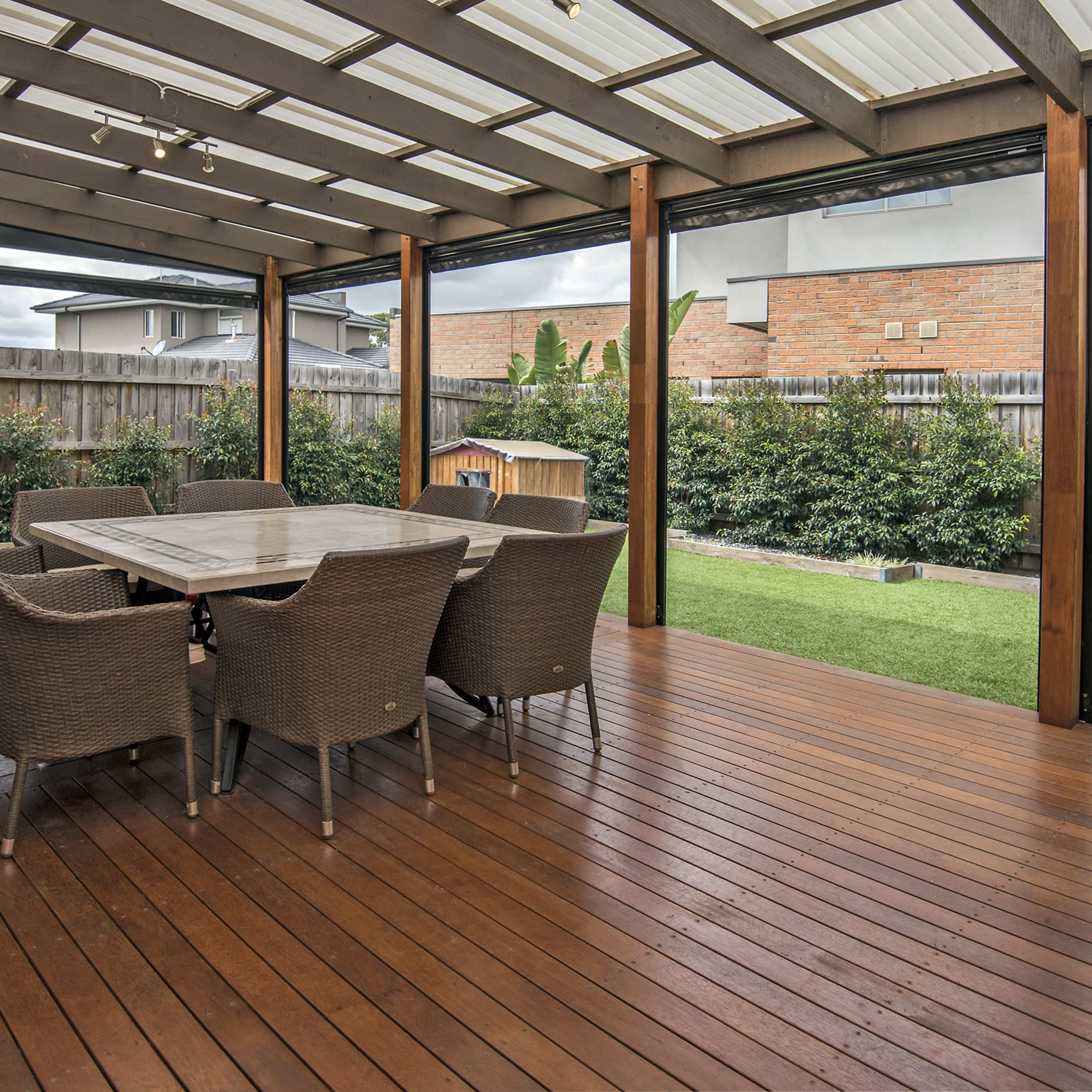 A covered outdoor patio with a wooden deck, featuring a large dining table surrounded by wicker chairs, overlooking a grassy yard and shrubs.