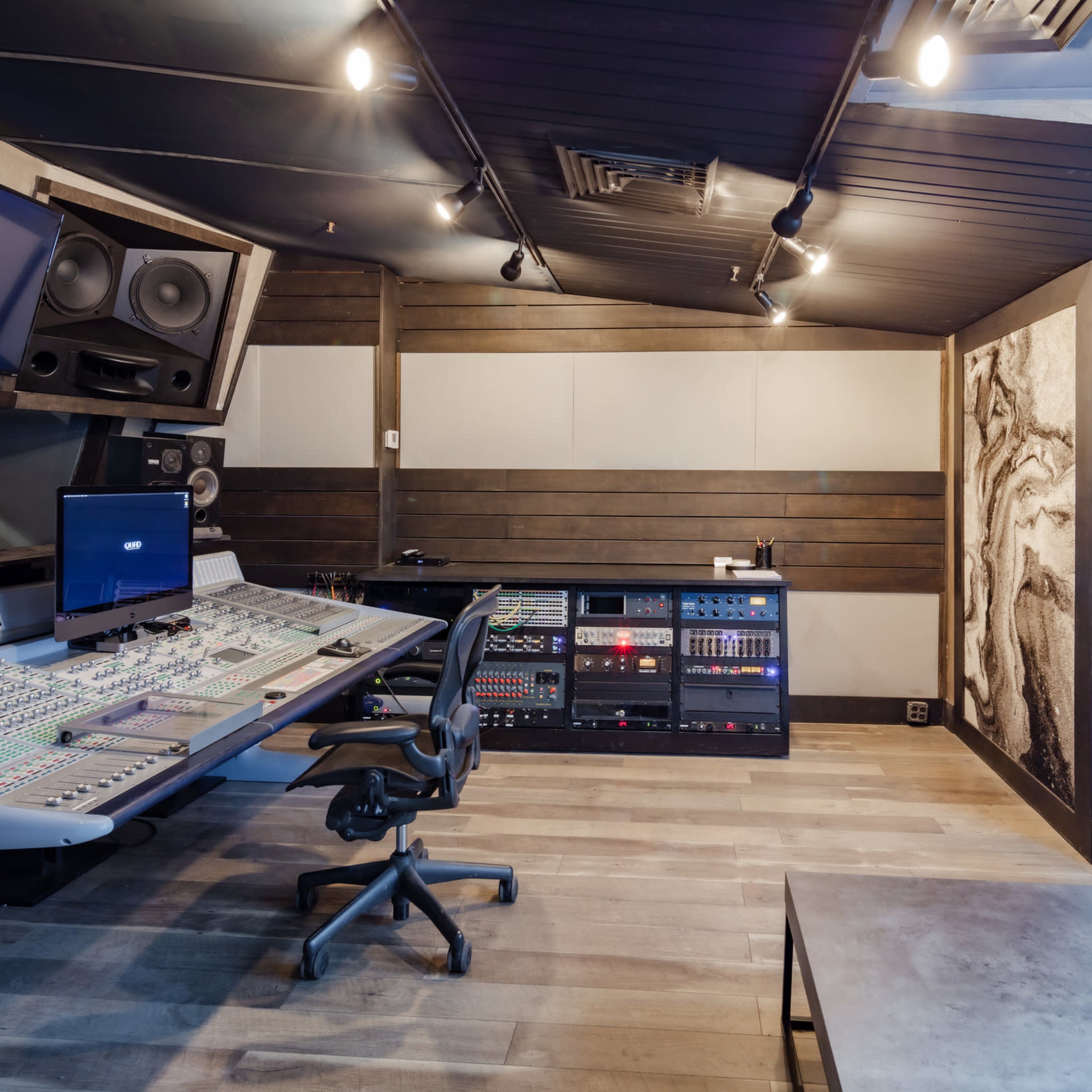 A music recording studio with a large mixing console, black seating, and acoustic wall panels.