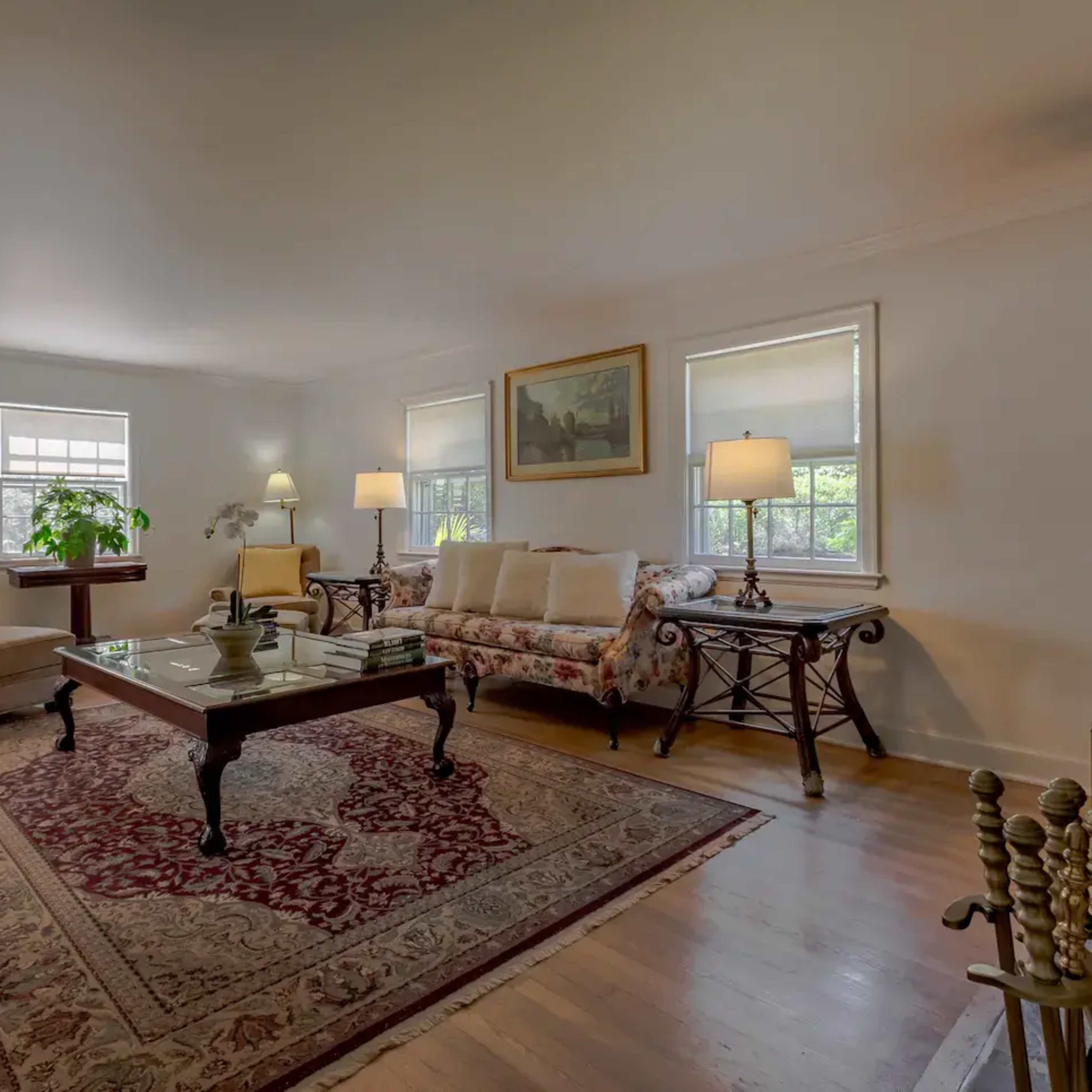 A living room featuring a floral-patterned couch, two armchairs, a wooden coffee table, and a rug, with natural light coming through two windows.