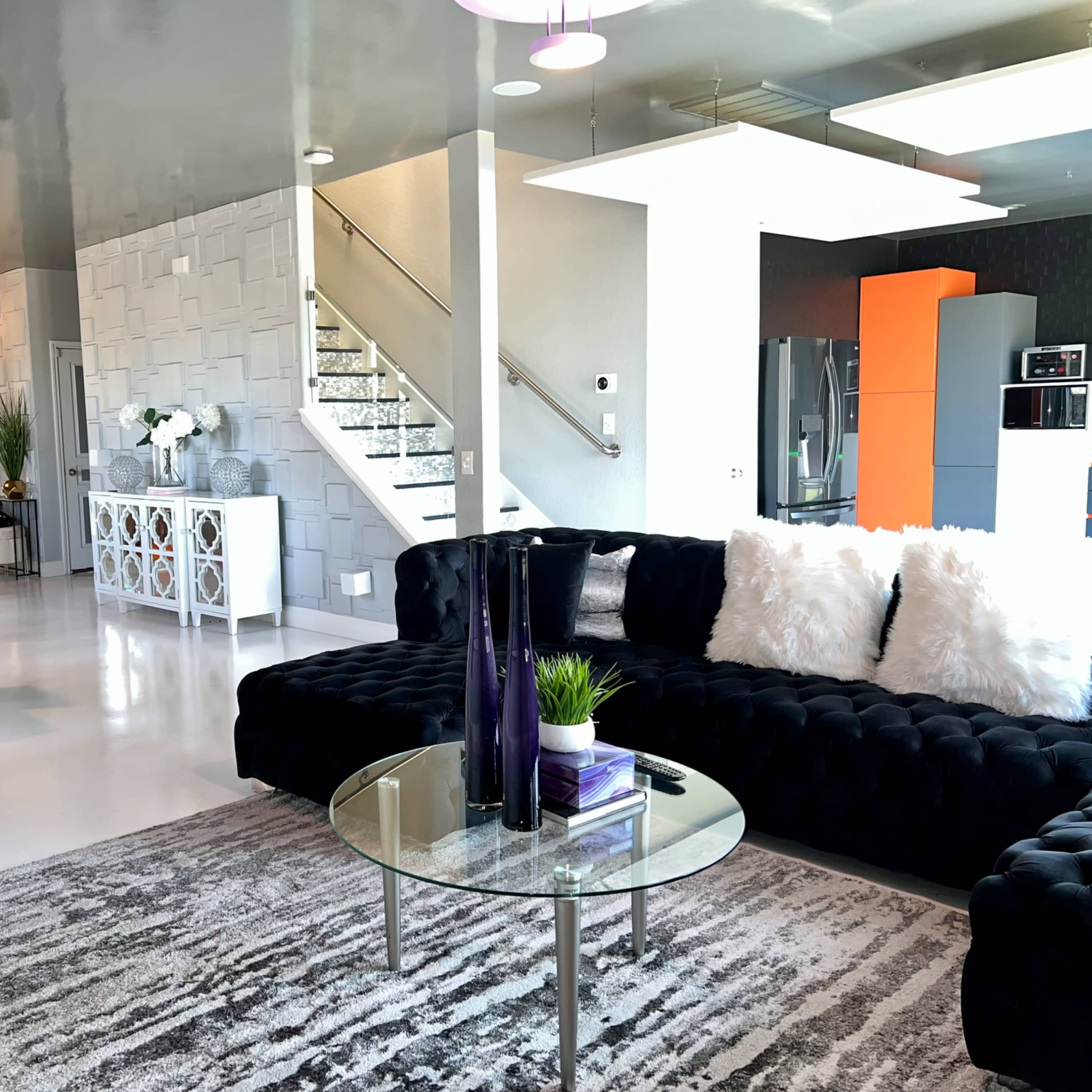 A modern living space featuring a black velvet sofa, a round glass coffee table, and an open dining area, with a staircase in the background.