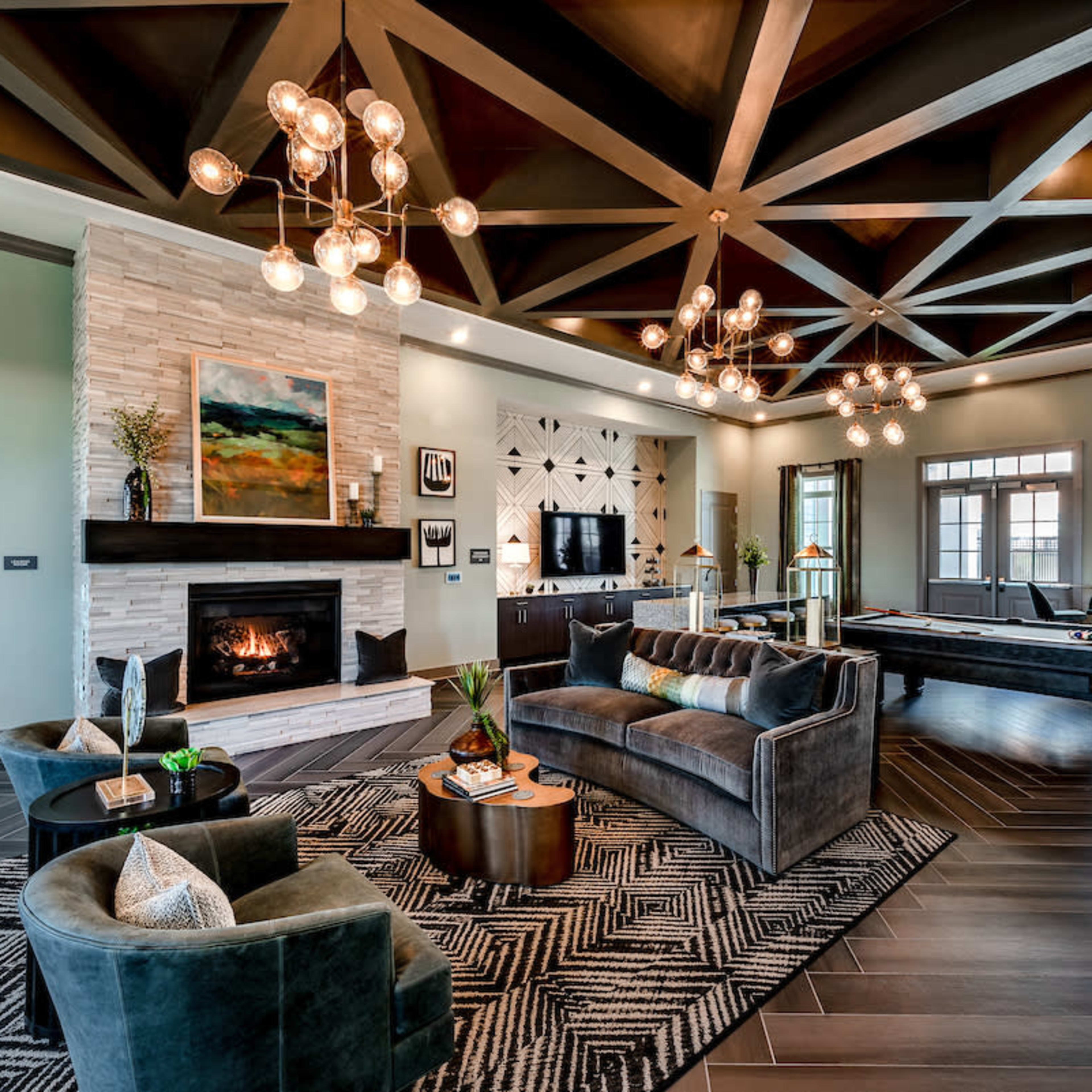 A modern living room featuring a stone fireplace, dark wood accents, large windows, and a pool table, all illuminated by decorative overhead lighting.