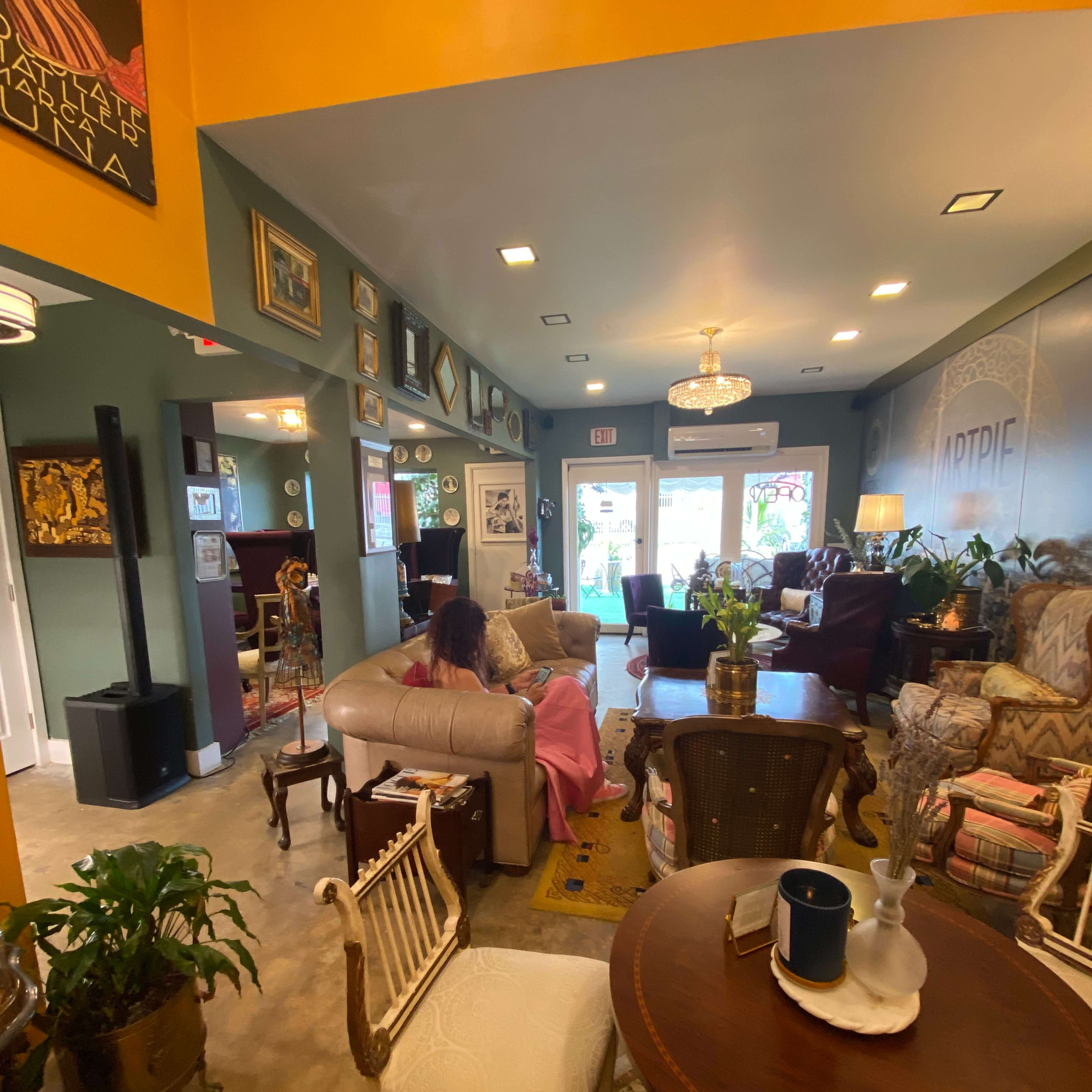 A cozy, eclectic interior of a café or lounge with various seating areas, decorative art on the walls, and large windows letting in natural light.