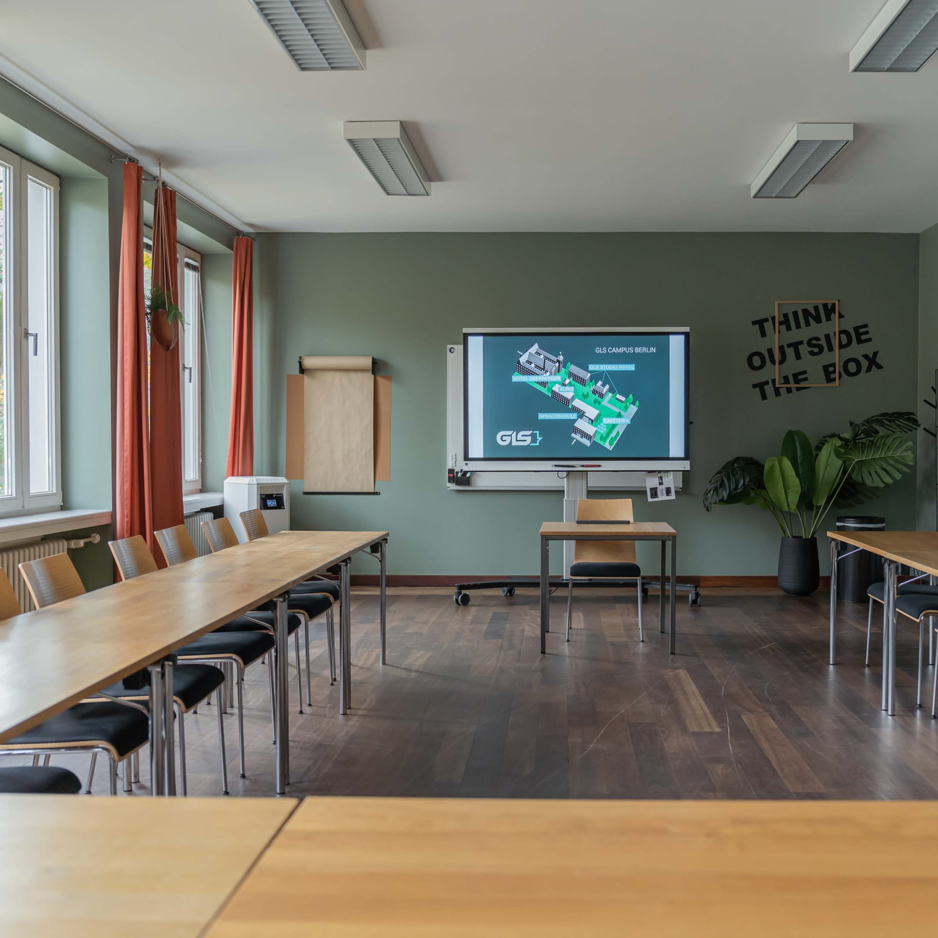 A conference room with a large table arranged in a U-shape, featuring a projector screen displaying a presentation, surrounded by wooden chairs and plants.