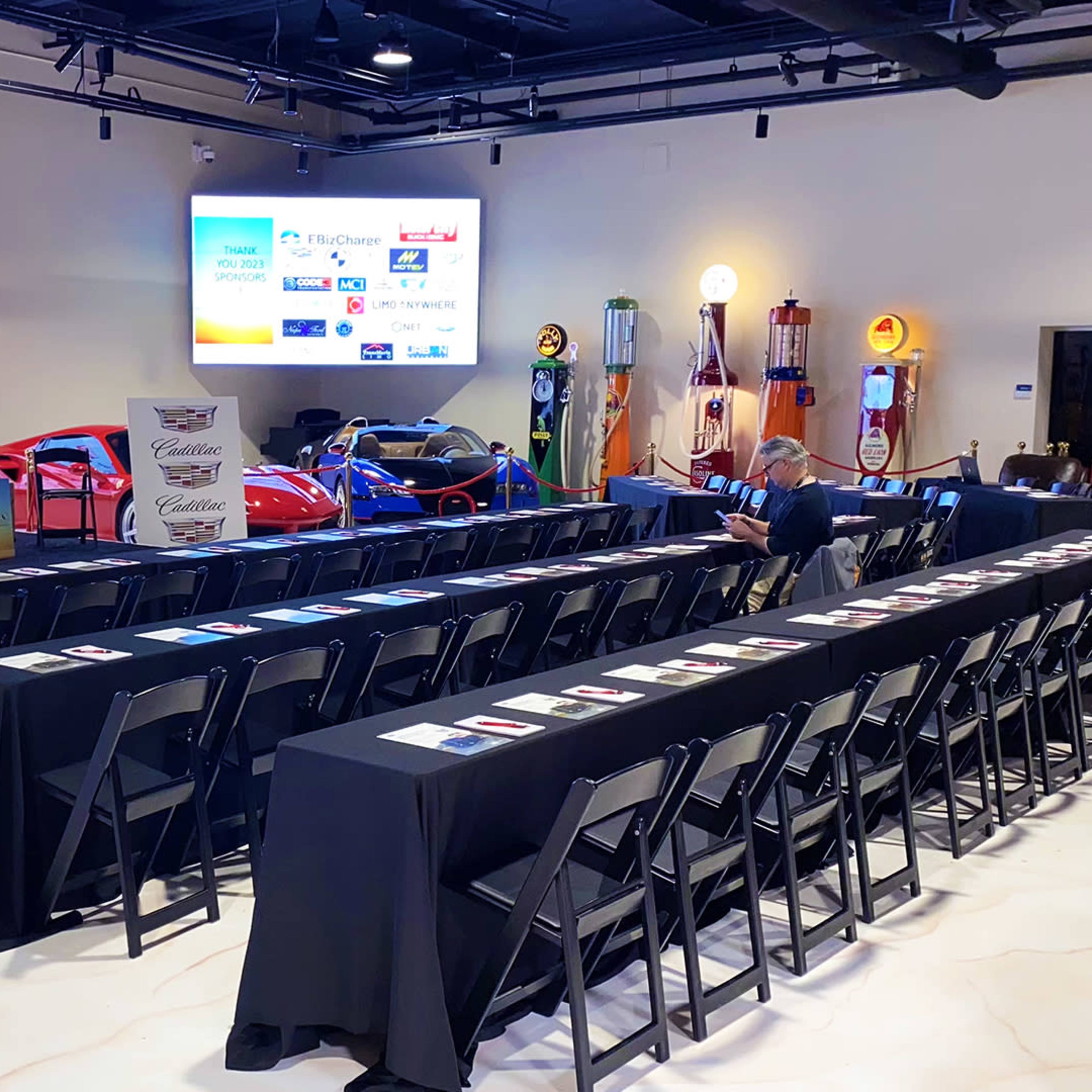 A spacious indoor event venue set up with black tables and chairs facing a presentation area, adorned with sponsor logos on large screens and vintage cars on display.