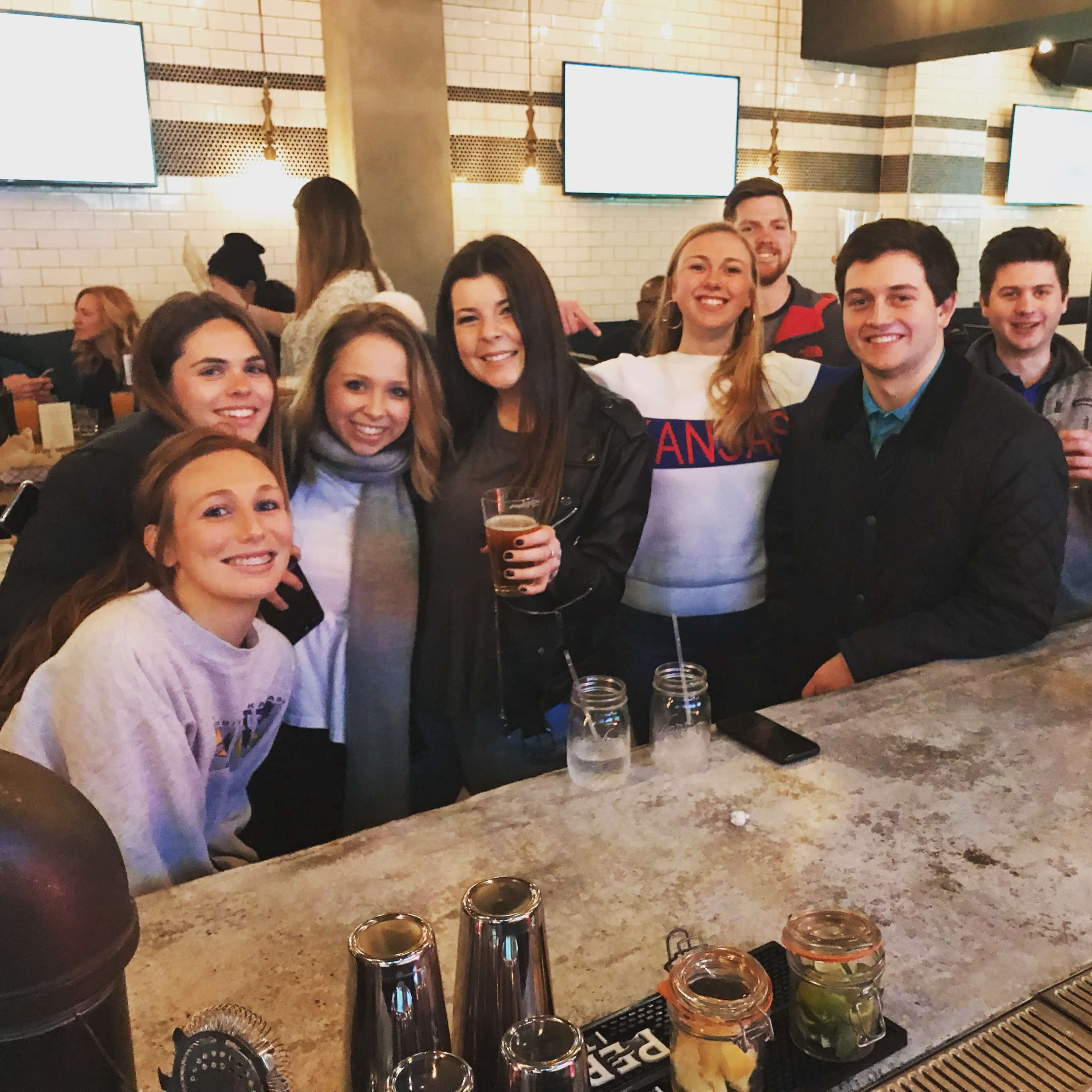 A group of nine people is gathered at a bar, smiling and holding drinks, with screens displaying content in the background.