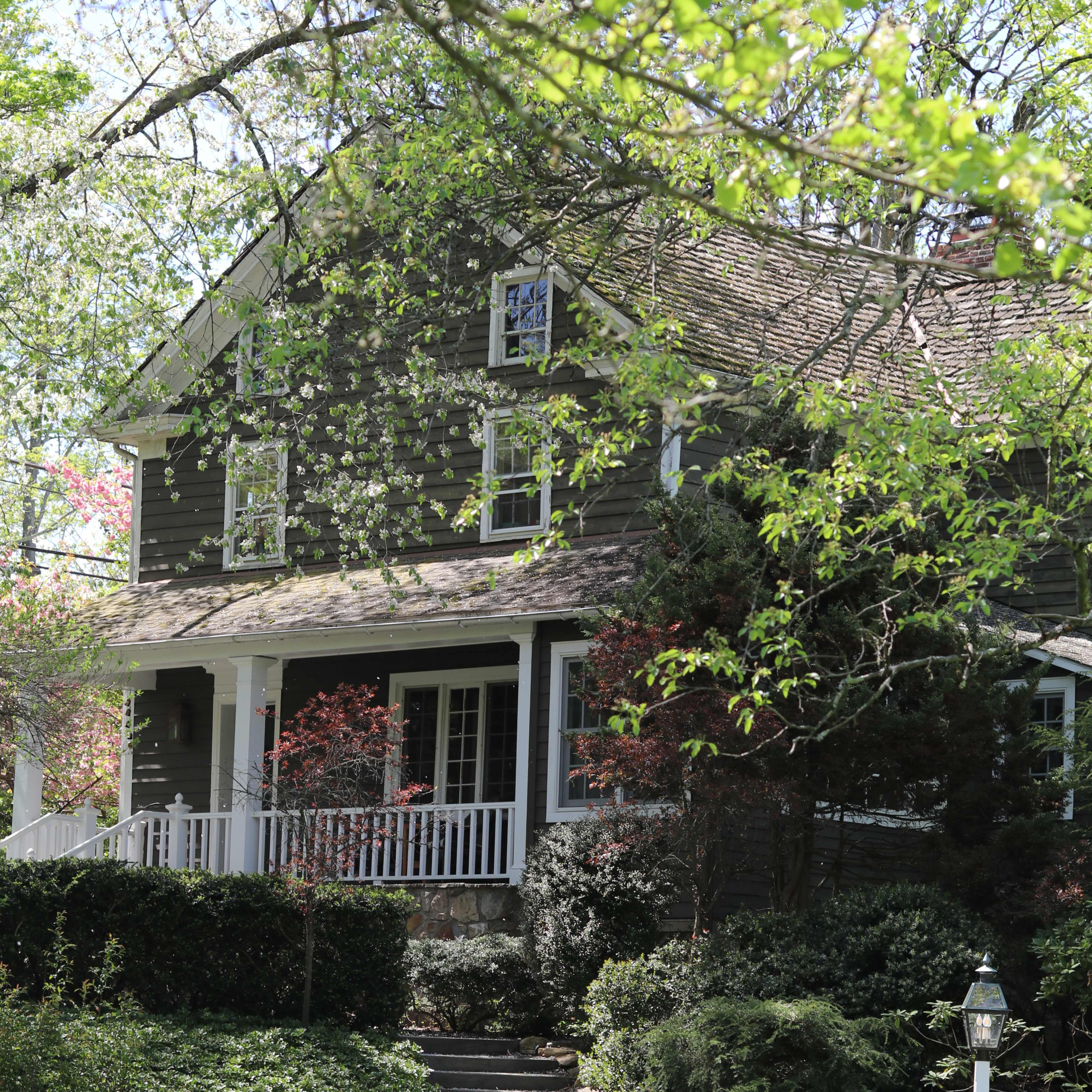 A two-story house with a front porch is surrounded by greenery and stone steps leading to the entrance.