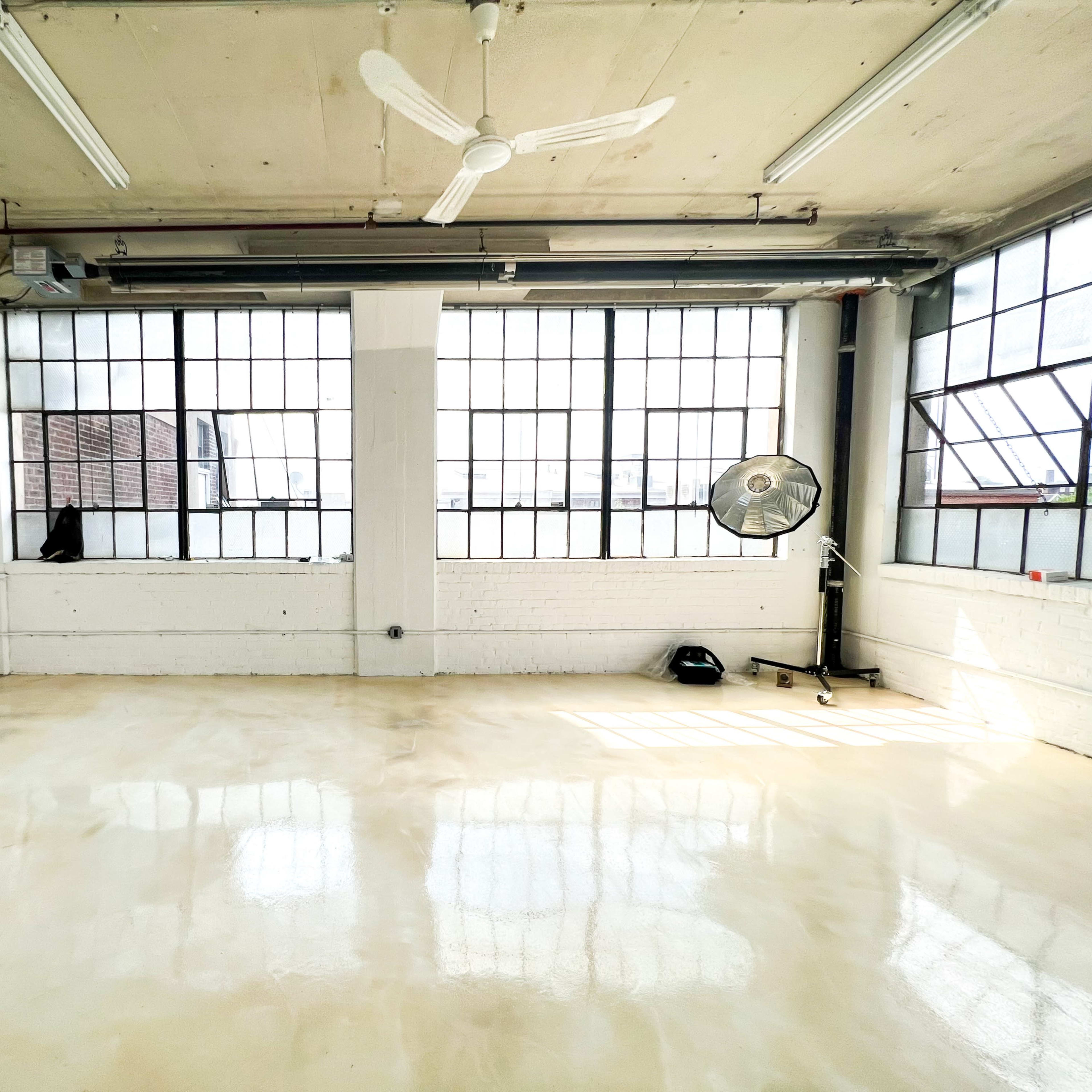 A spacious room with large industrial windows, a ceiling fan, and a polished floor.