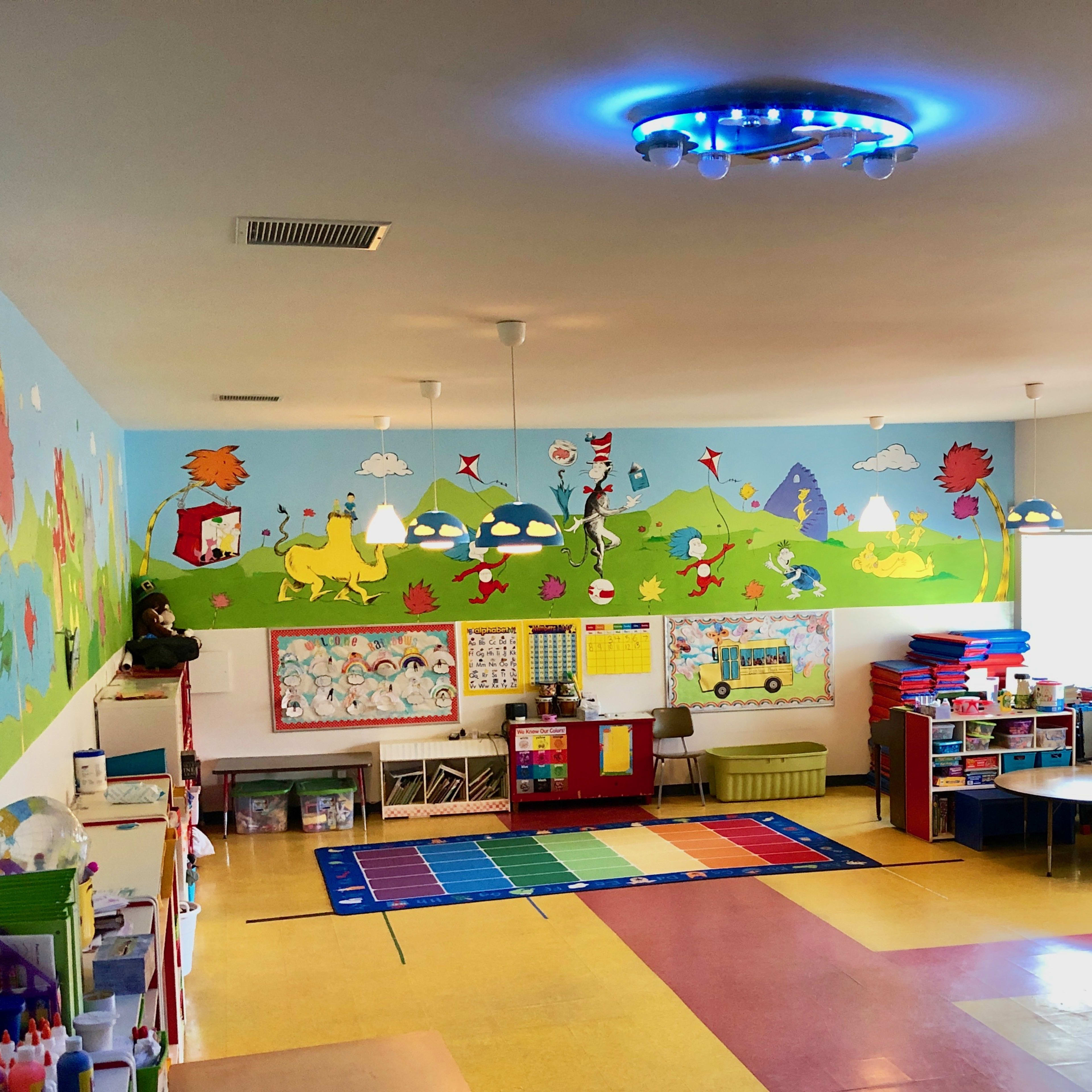 A brightly colored classroom features a mural of animals and landscapes, with tables and storage areas for educational materials.