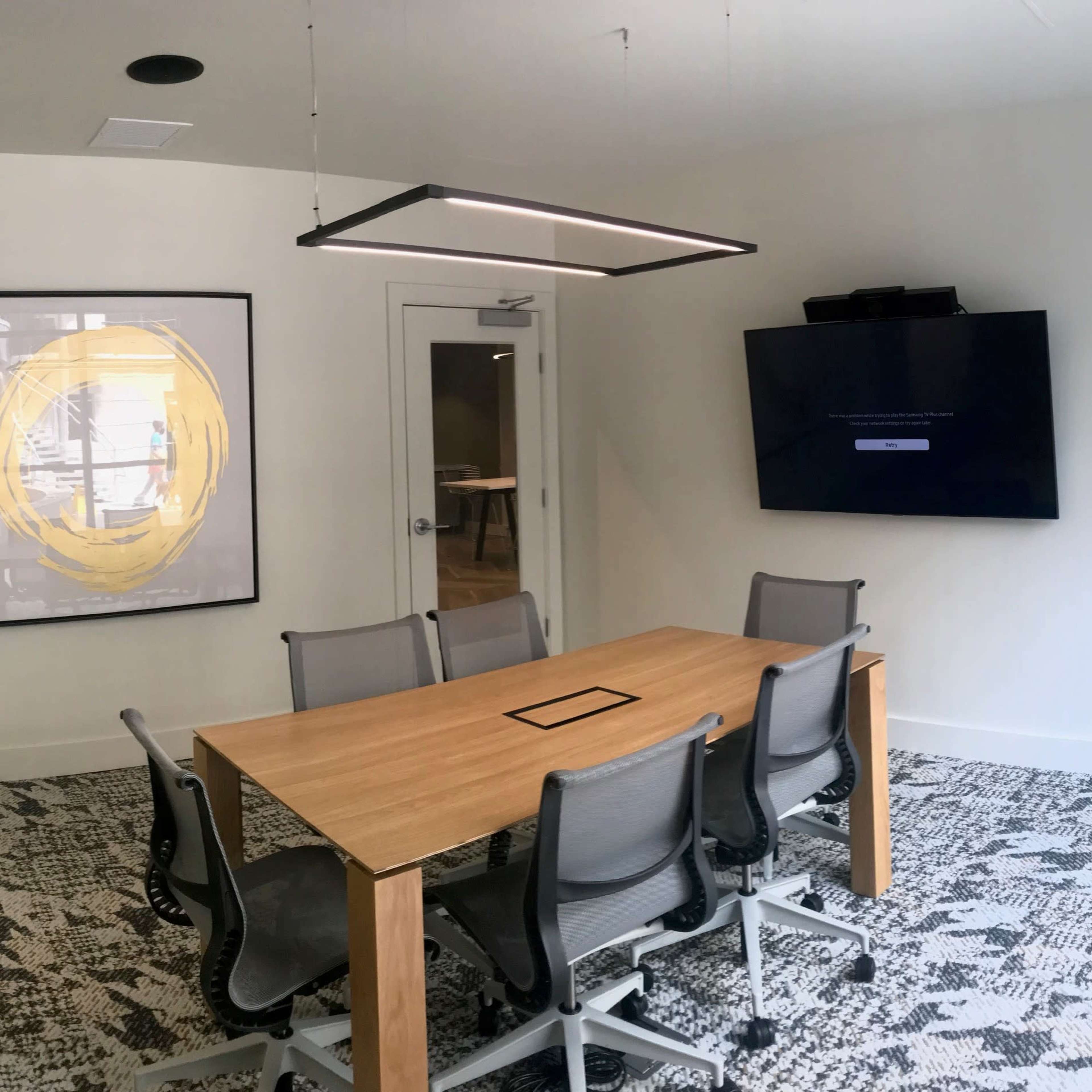 A conference room features a wooden table surrounded by six chairs, a wall-mounted screen, and a large framed artwork.