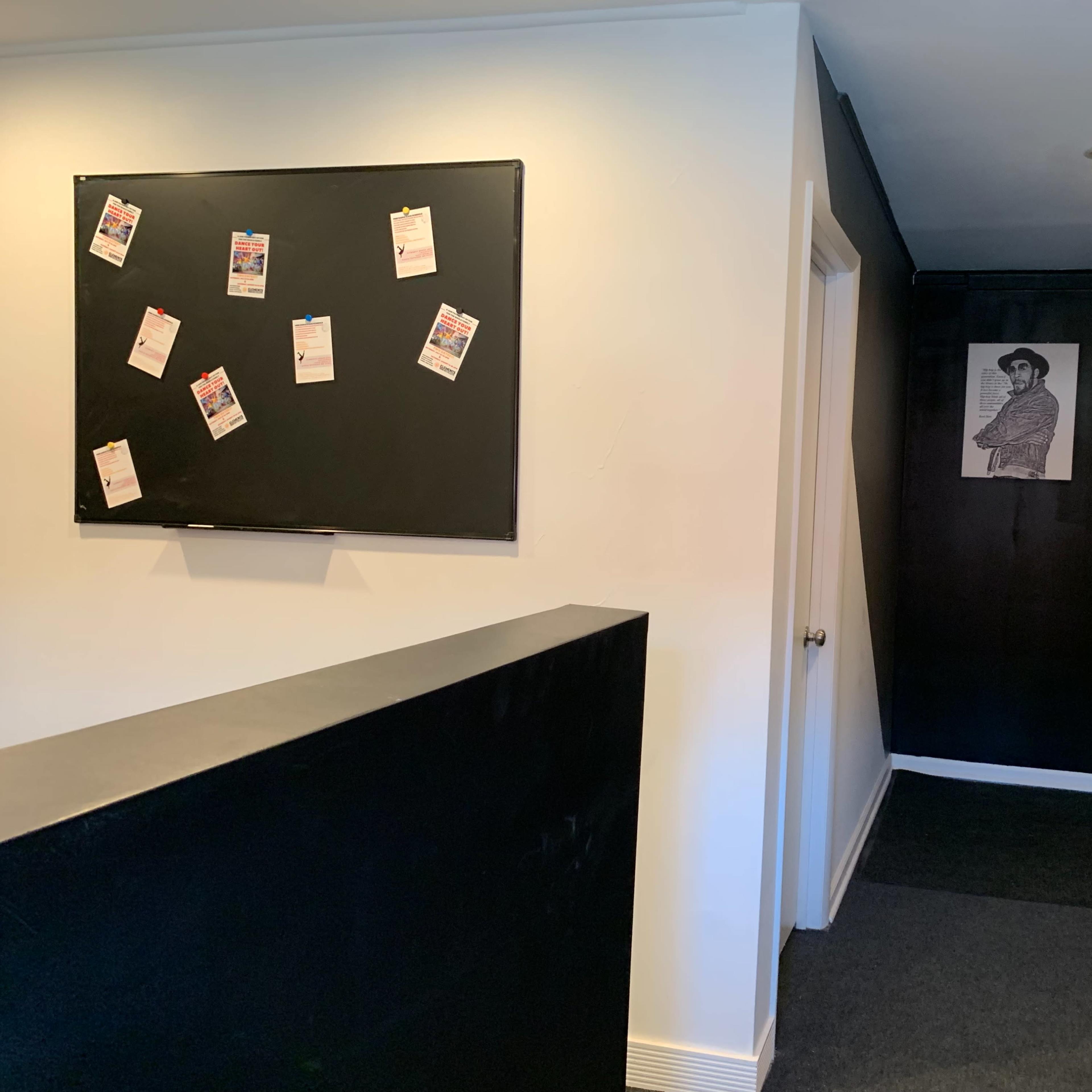 A hallway with a black bulletin board on the left, a dark wall on the right, and a doorway at the end of the corridor.