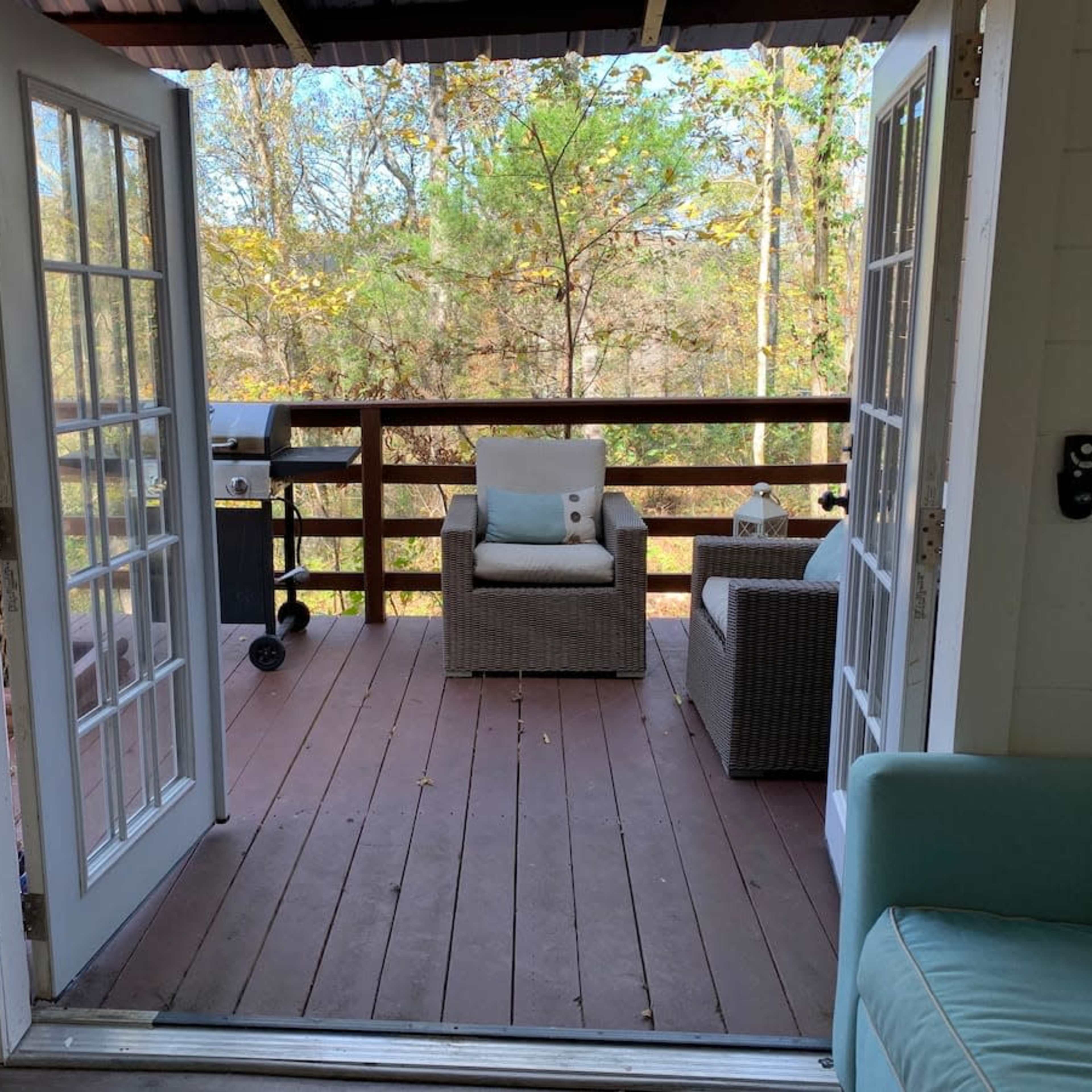 A balcony with two chairs, a small table, and a grill, framed by open French doors that lead to a wooded area.