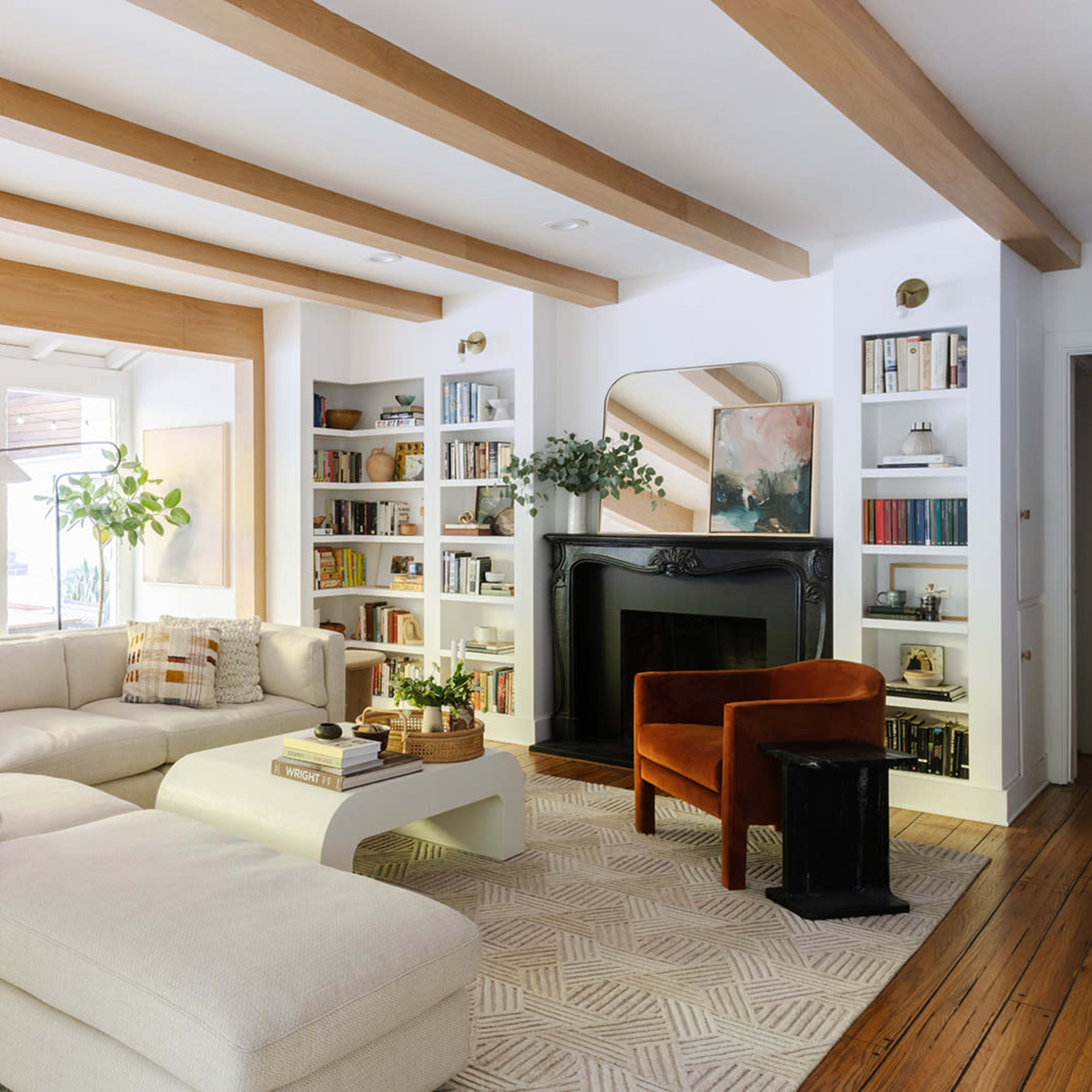 A spacious, well-lit living room featuring a light-colored sectional sofa, a fireplace, bookshelves, and large windows that open to a garden.