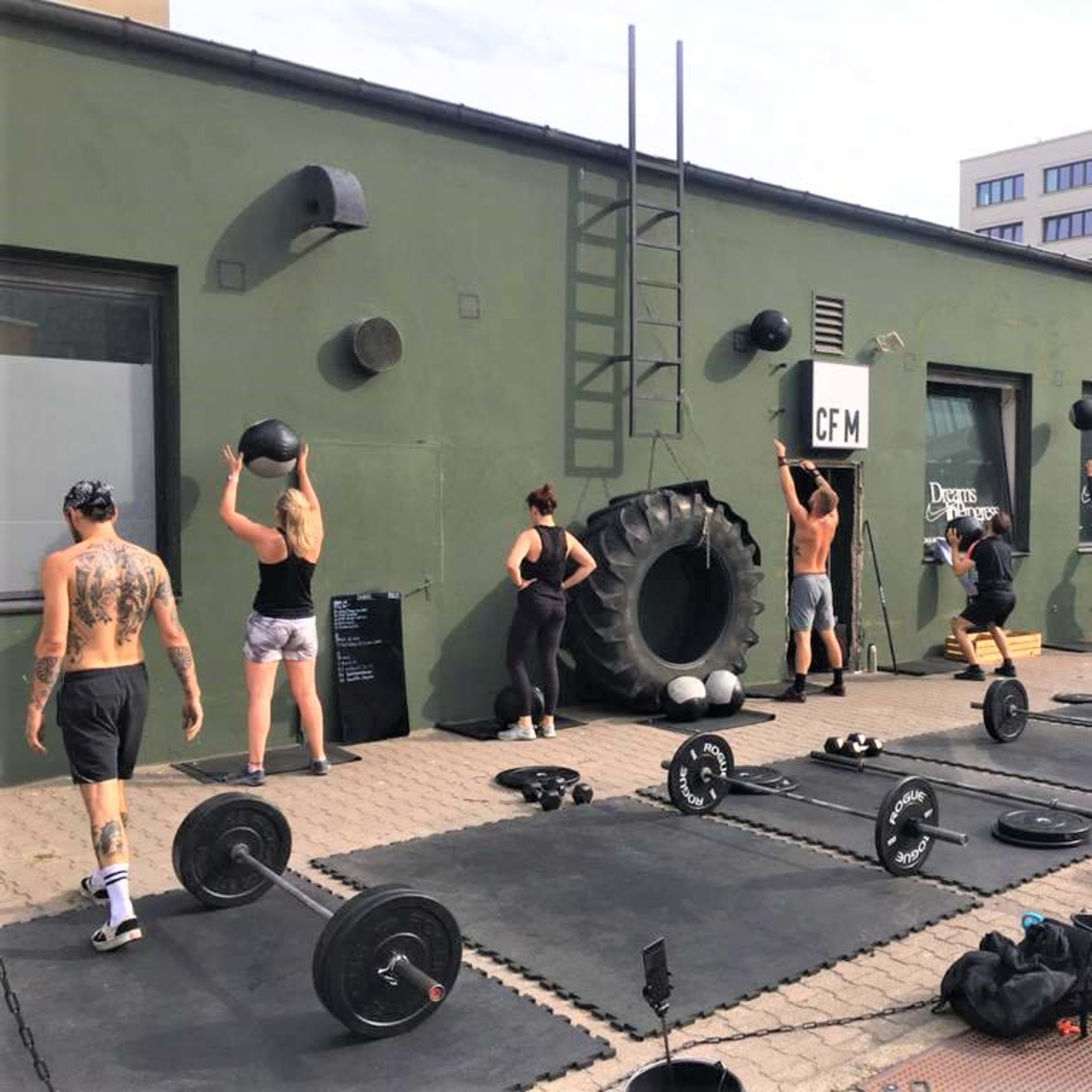 A group of people are participating in a workout session outside, using weights and medicine balls near a gym with a large tire and exercise mats.
