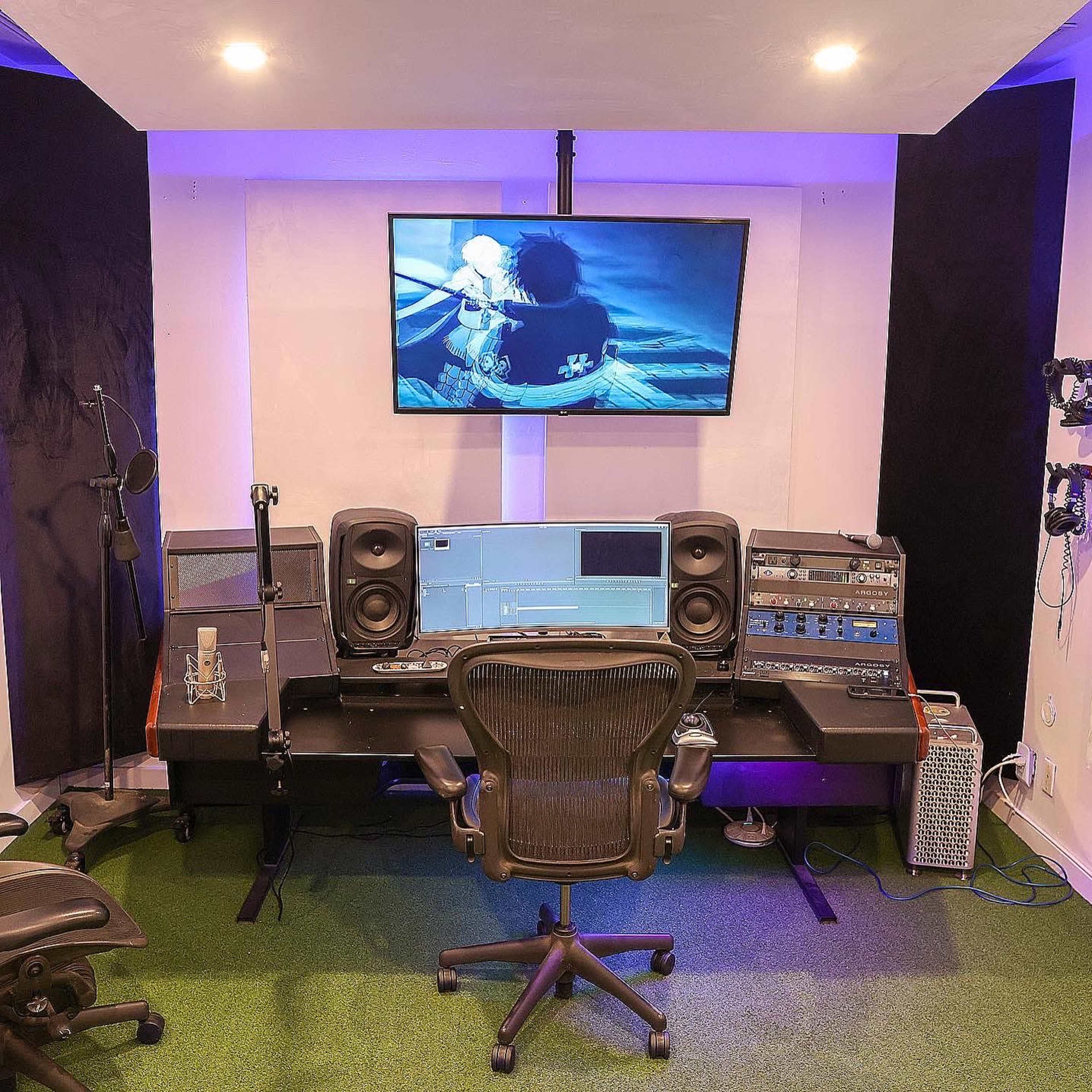 A sound editing workspace with a desk, audio equipment, a computer monitor displaying a video, and green carpet flooring.