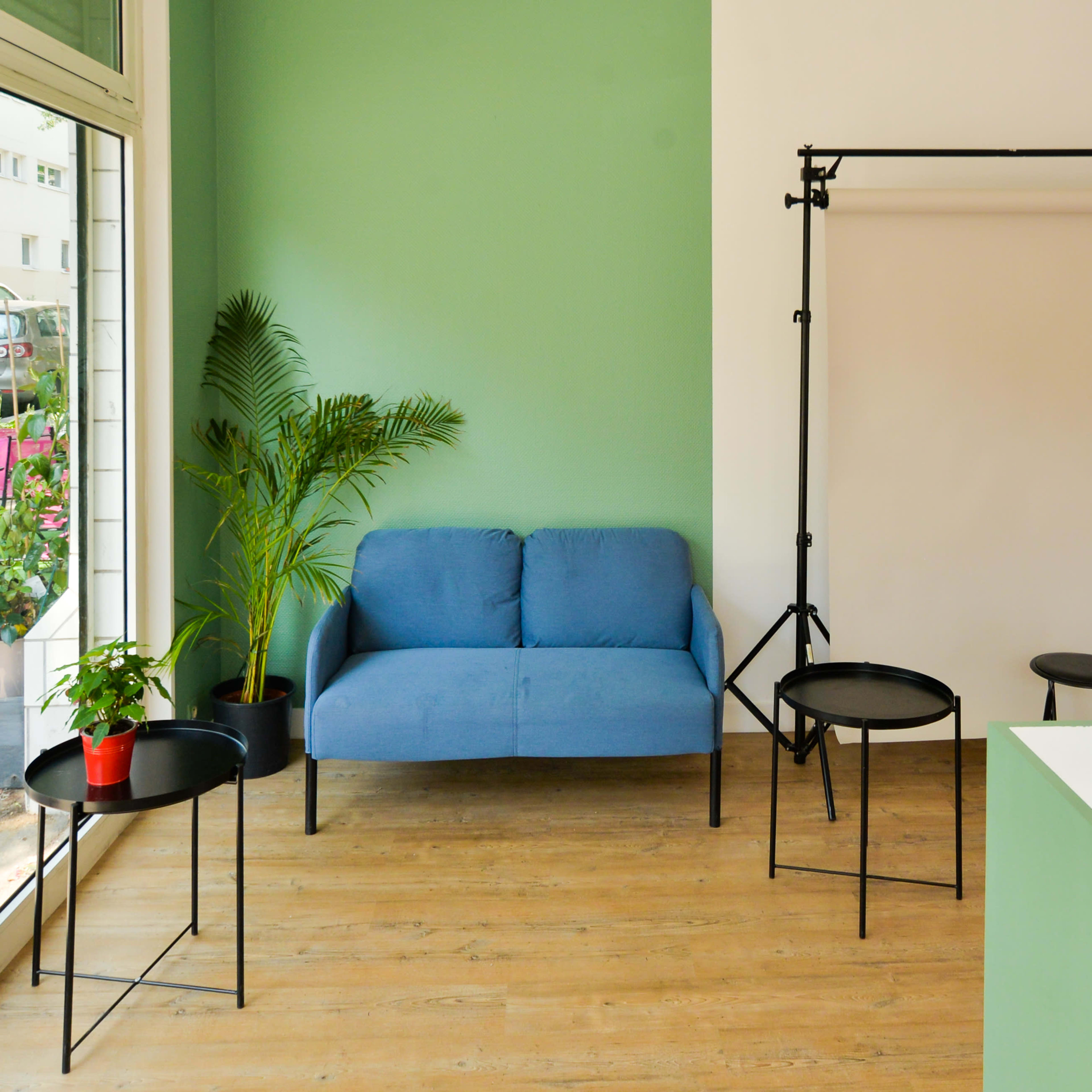 A minimalistic interior space featuring a blue couch, black side tables, a stool, and a potted plant against a green wall.