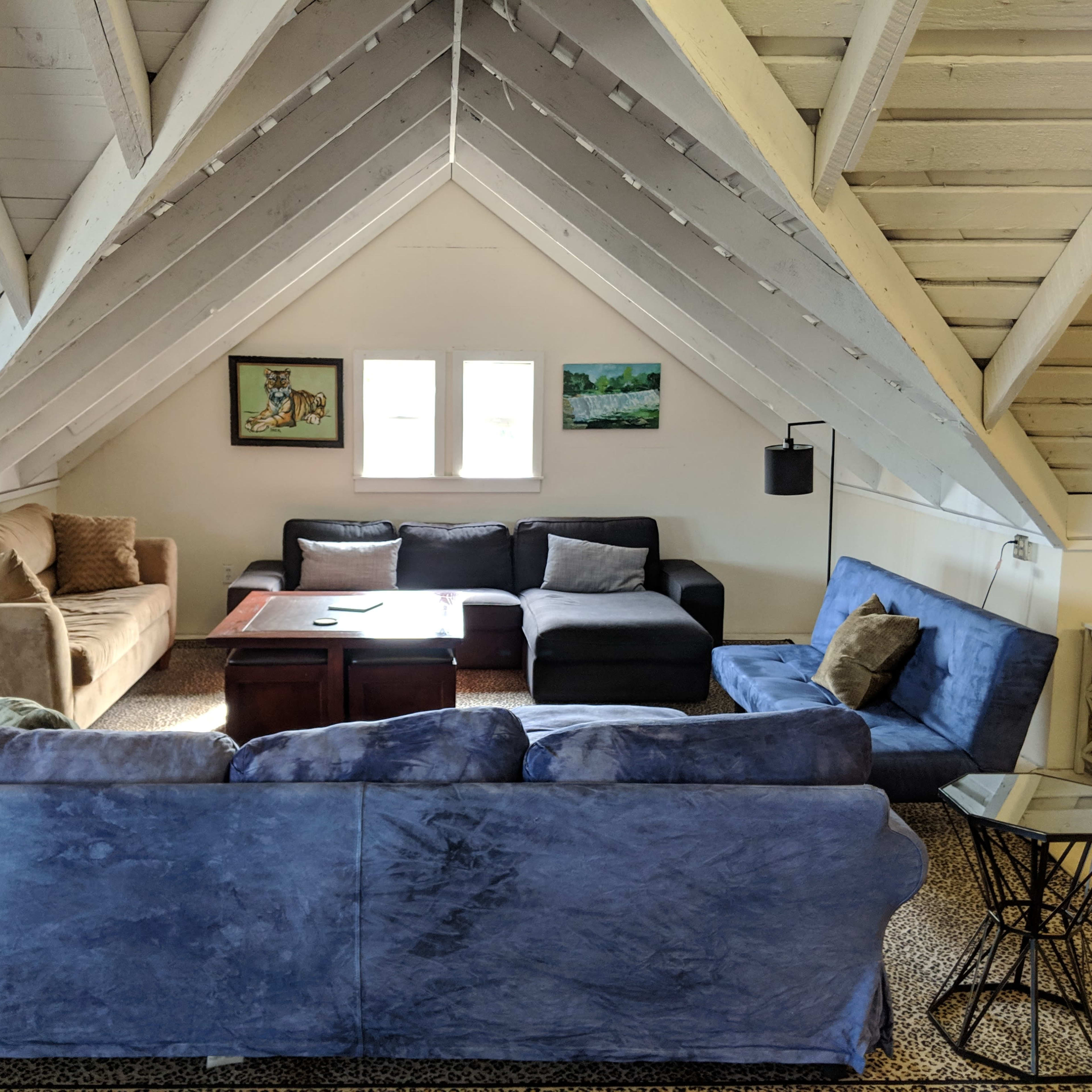 The scene features a spacious, sloped-ceiling living area with several sofas, a coffee table, and large windows allowing natural light.