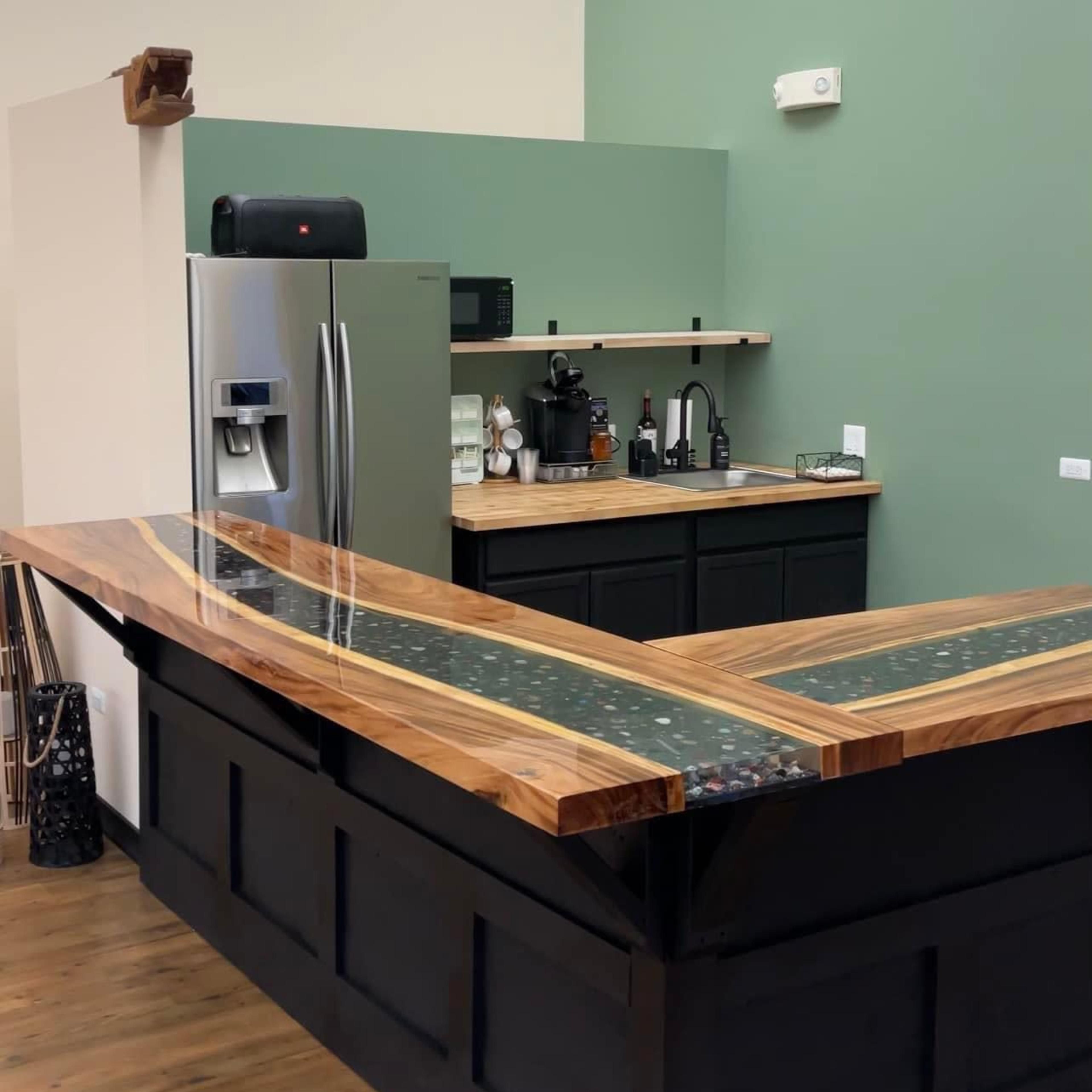 A modern bar area featuring a sleek wooden countertop with embedded stones, a refrigerator, a sink, and a black cabinetry against a muted green wall.