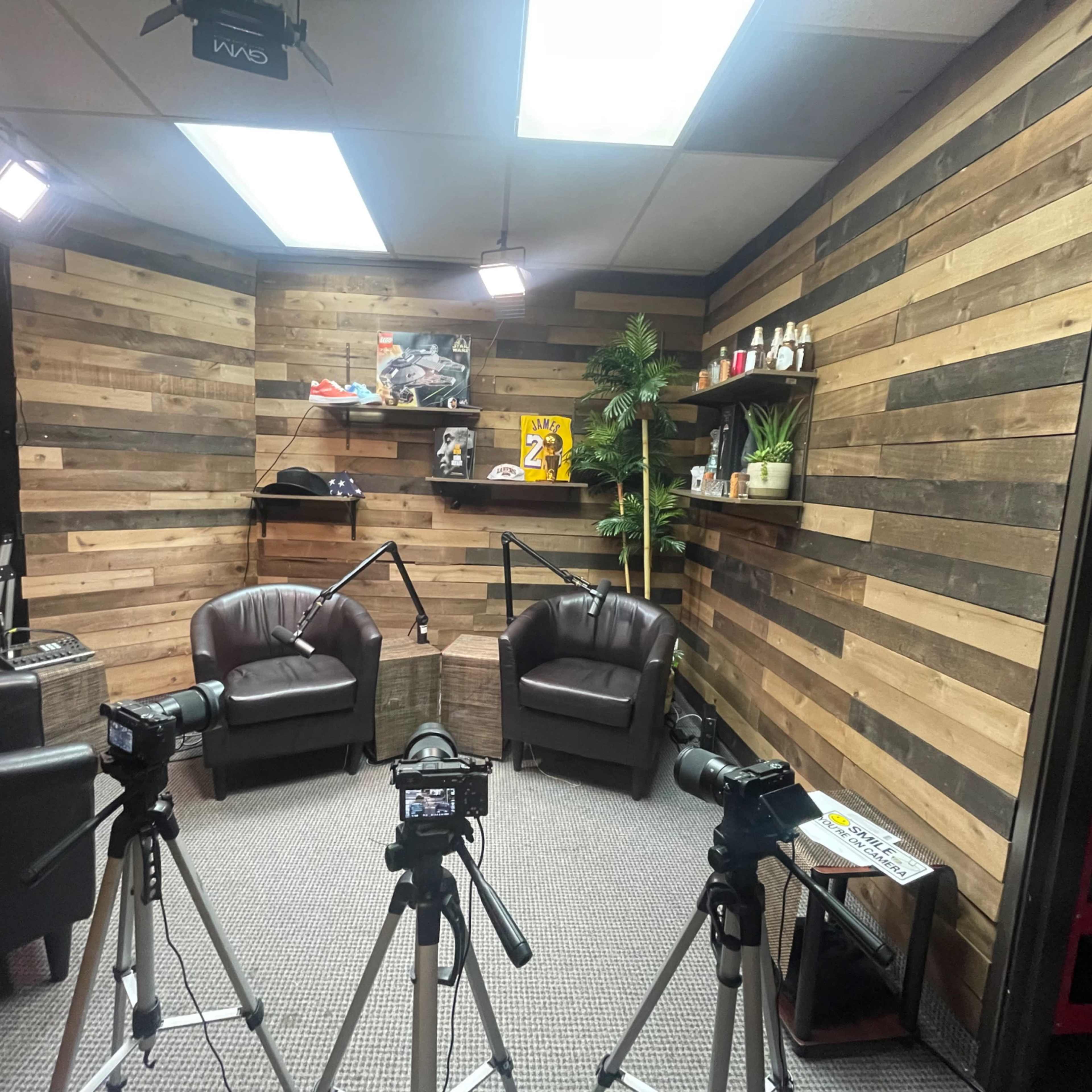 A recording setup in a room with wooden walls, featuring two black chairs, three cameras on tripods, and various decorative items on shelves.