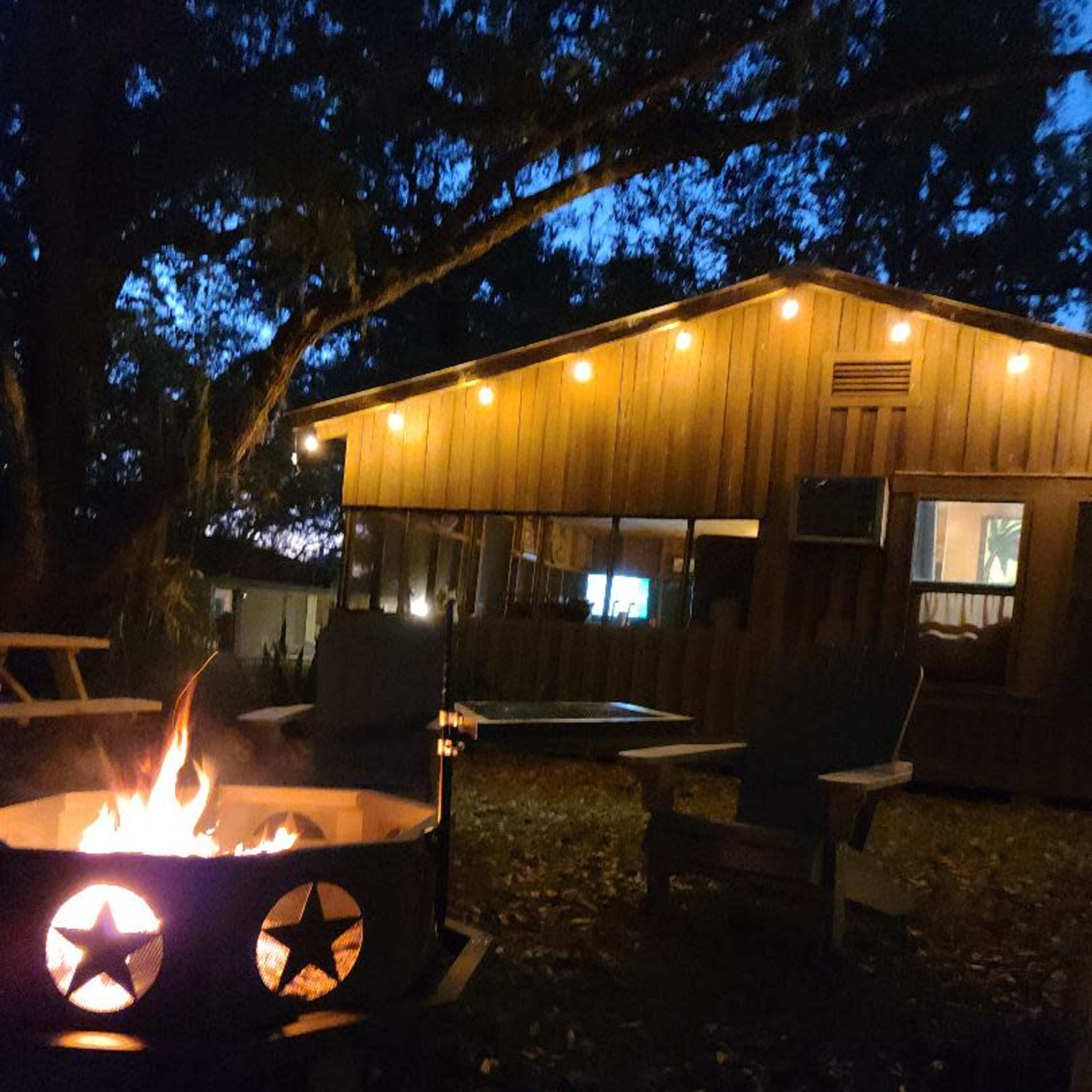 A fire pit with stars cut out burns in the foreground, while a wooden building illuminated by indoor lights stands in the background under a twilight sky.