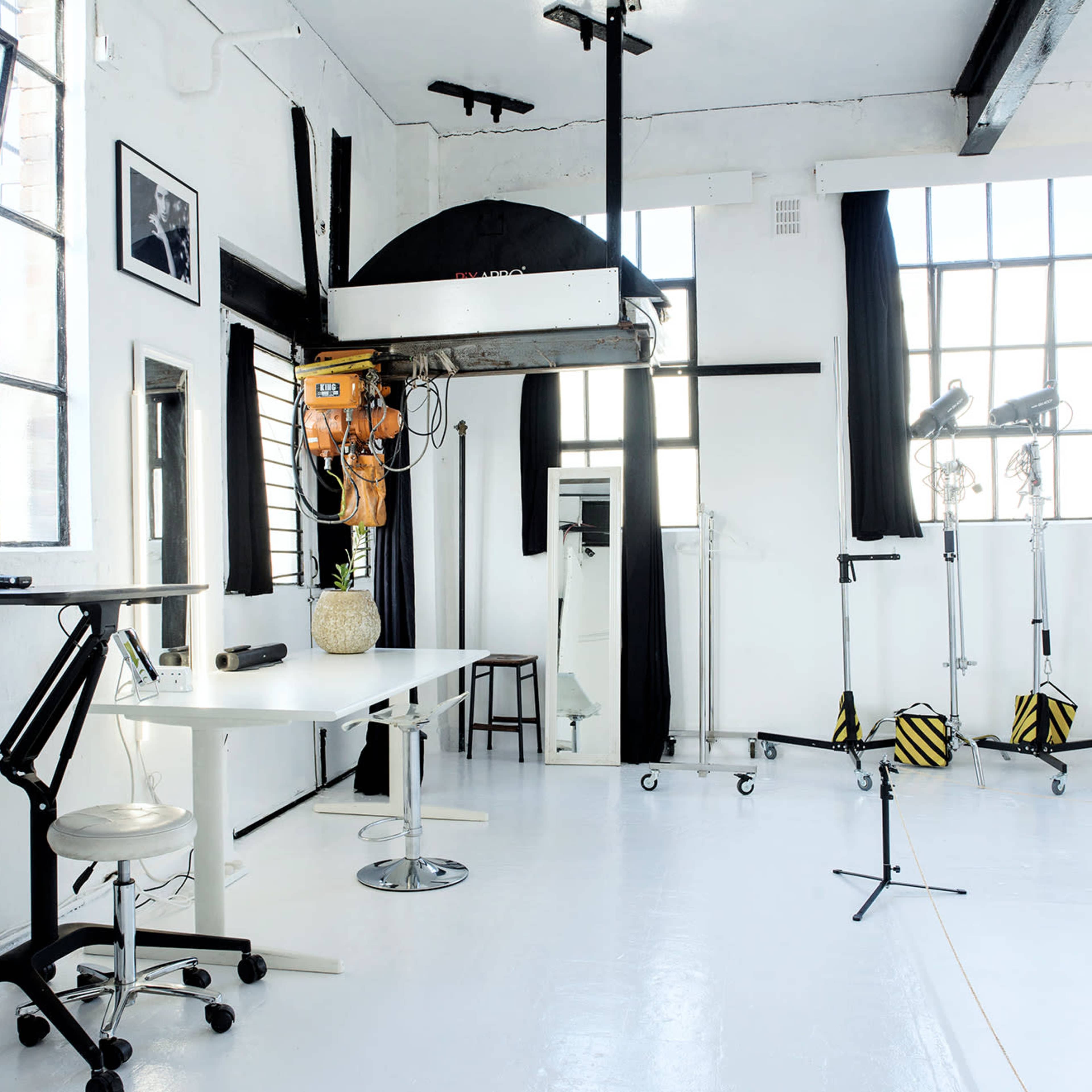 A bright, minimalist studio space with large windows, a table, a chair, and various lighting equipment.