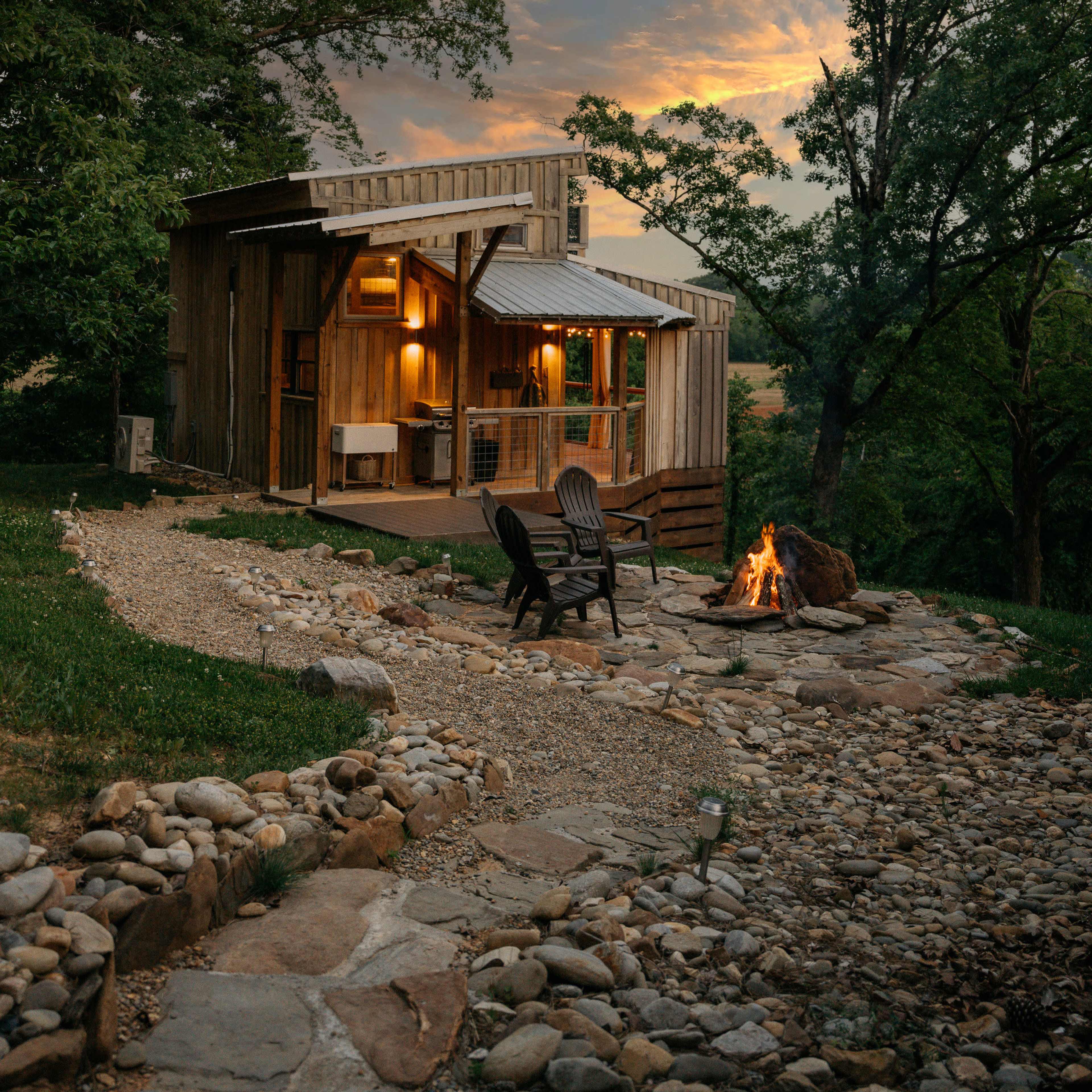 A wooden cabin with a stone path and fire pit is set against a sunset sky, surrounded by trees.