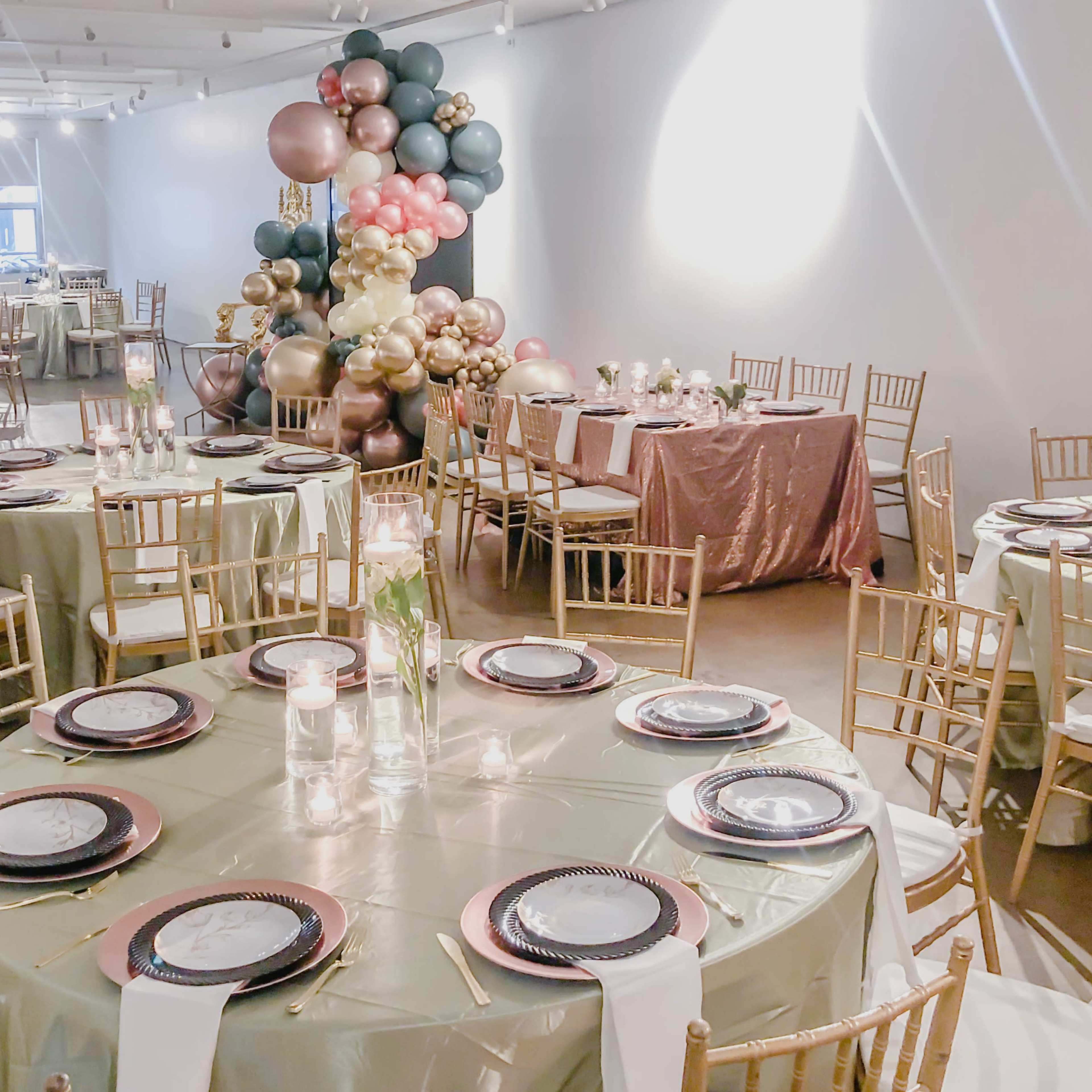 A spacious event venue set up with elegantly arranged tables, each featuring gold chiavari chairs, plates, and floral centerpieces, alongside a colorful balloon display in the corner.
