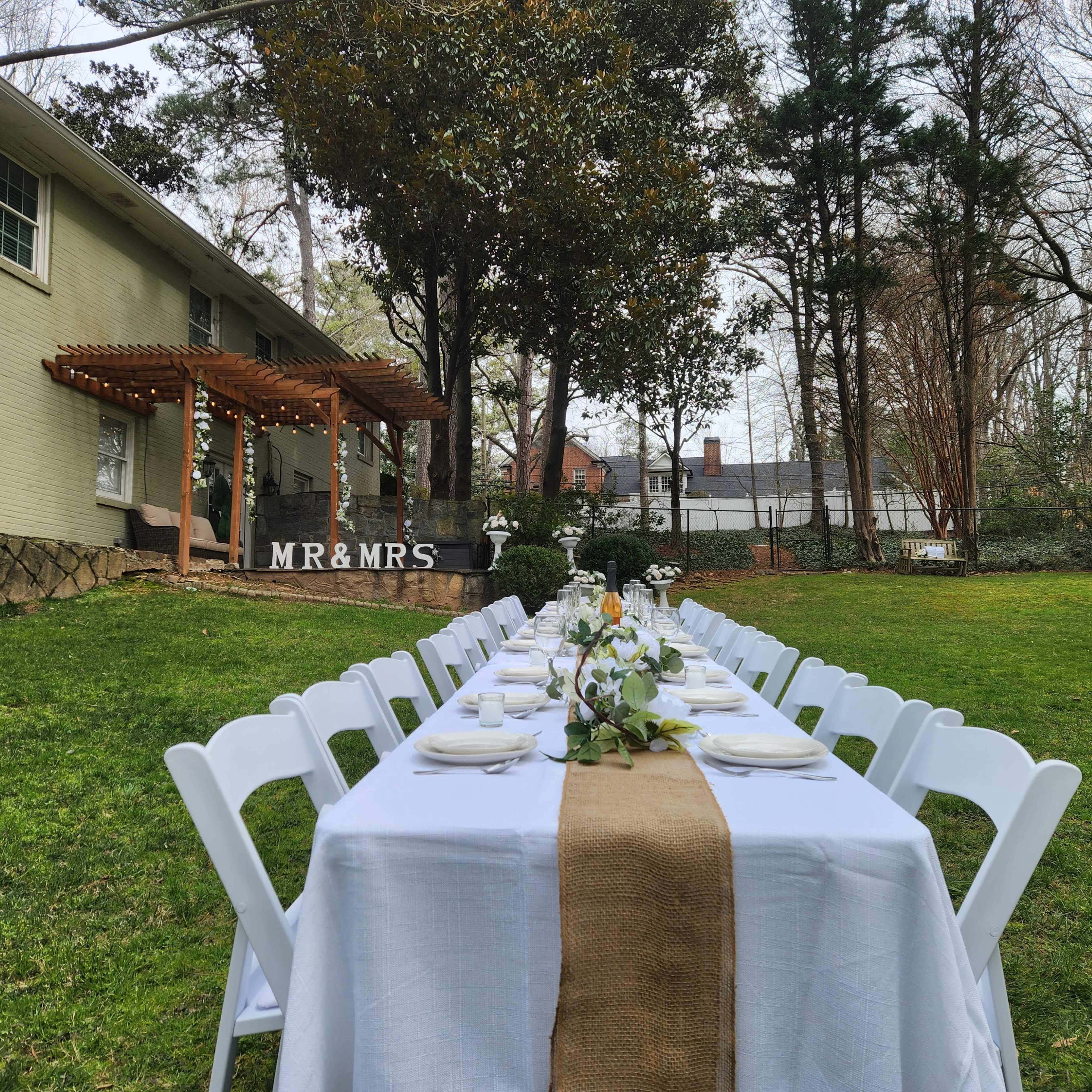 A long table set with white tablecloths and decor is arranged on a grassy area outside a house, with a wooden pergola and a sign reading "MR & MRS" in the background.