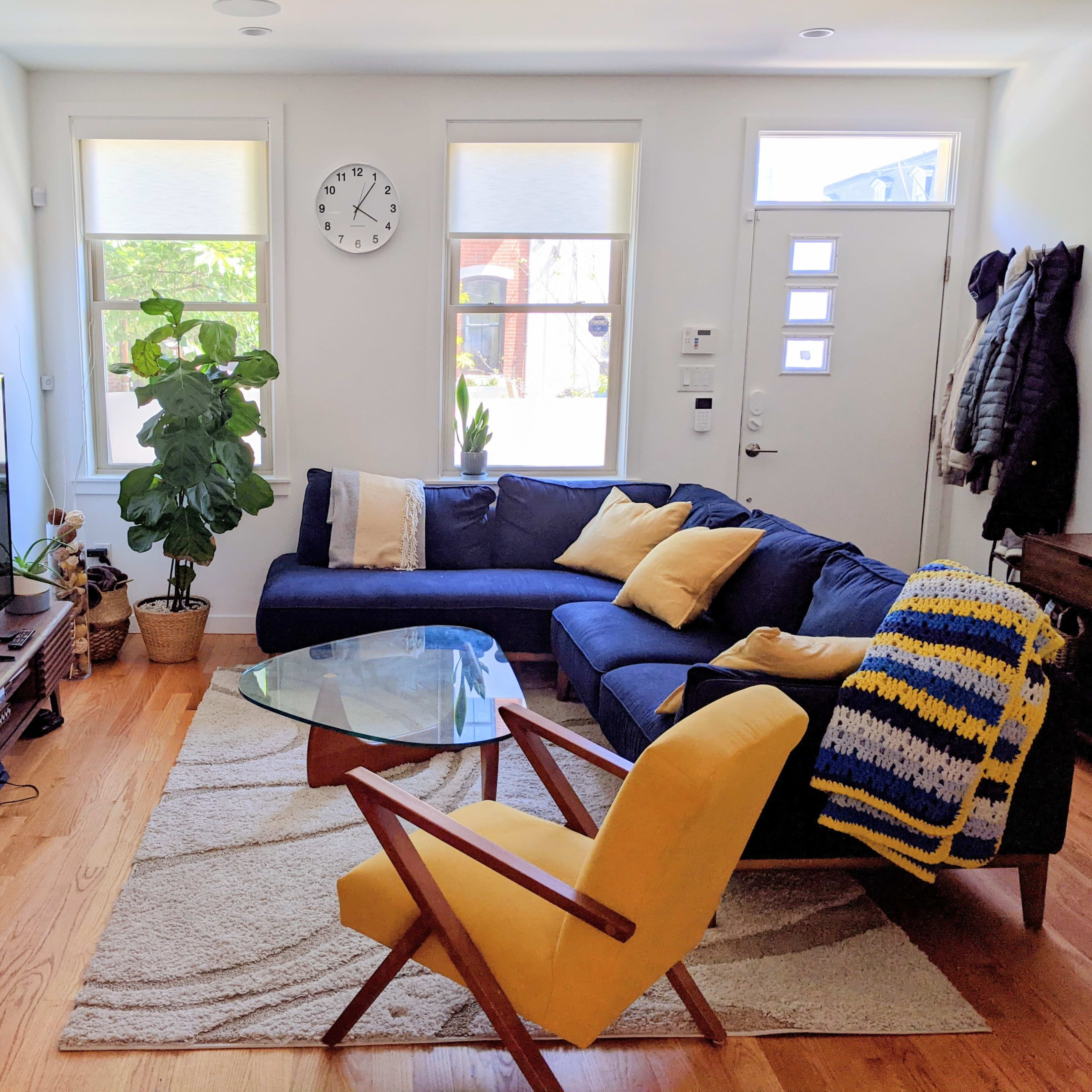 A living room featuring a blue sectional sofa, a glass coffee table, a television, and a yellow armchair, along with plants and a wall clock.