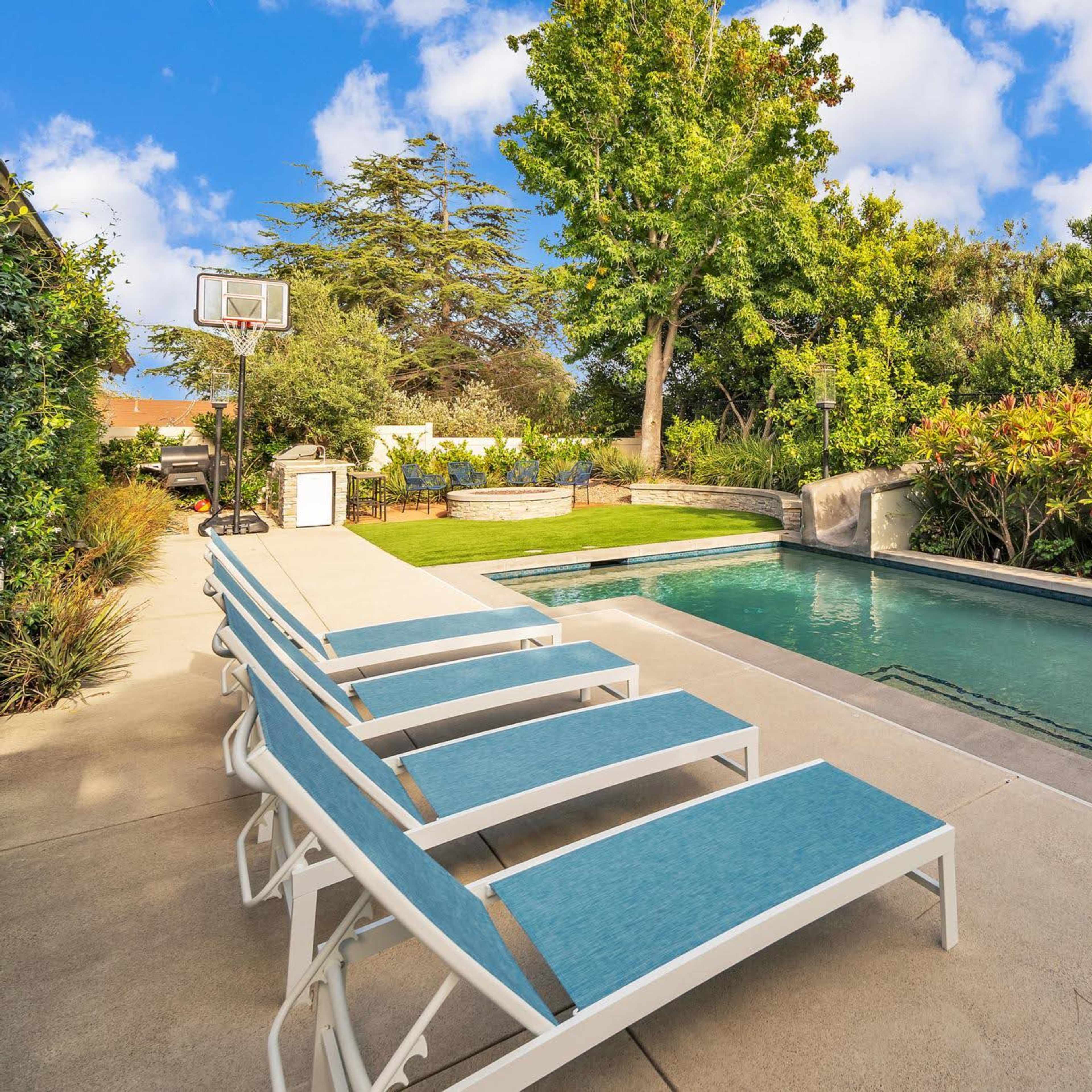 A backyard with a swimming pool surrounded by lounge chairs, landscaped greenery, and a basketball hoop in the background.