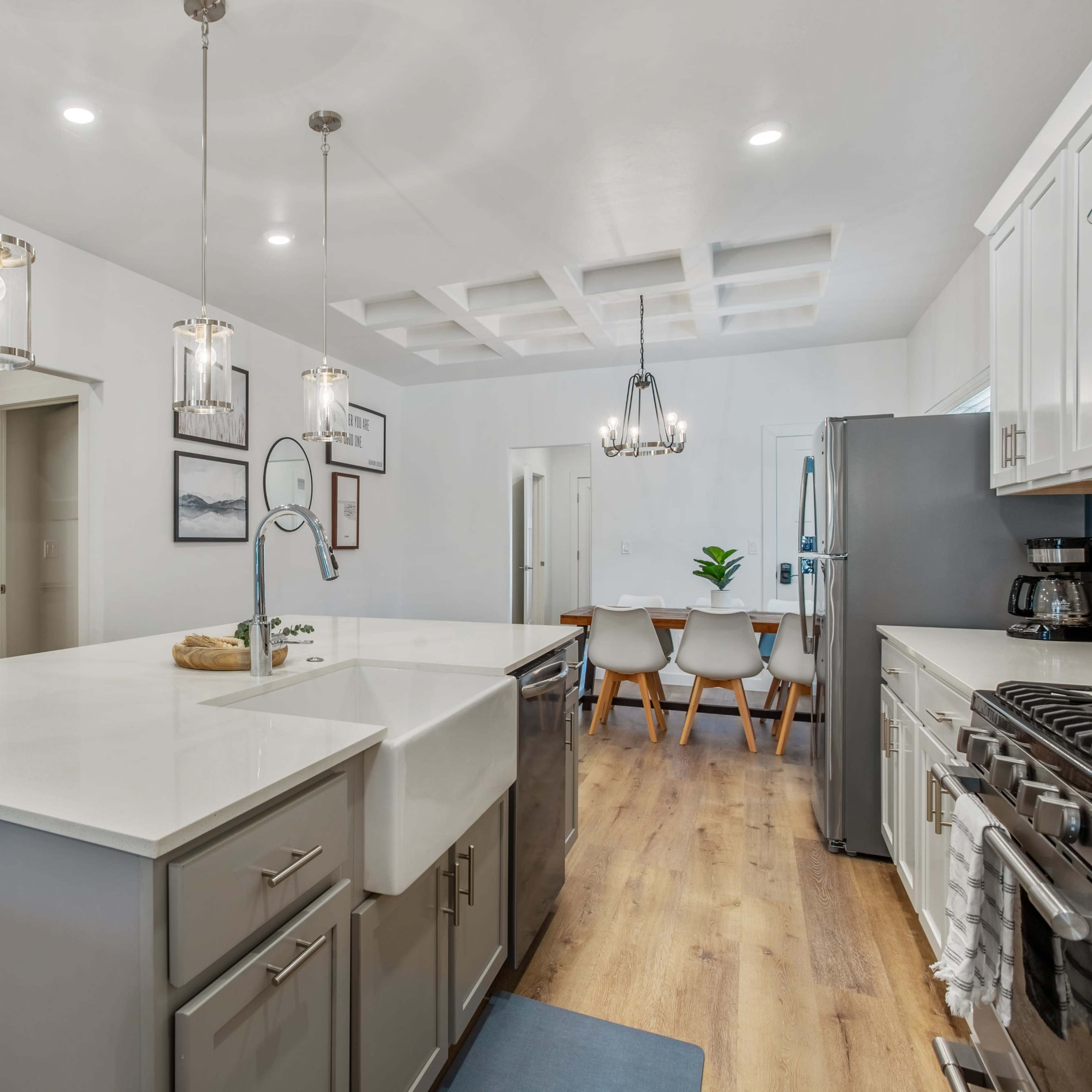 A modern kitchen features a large island with a farmhouse sink, pendant lighting, and stainless steel appliances, with a dining area visible in the background.