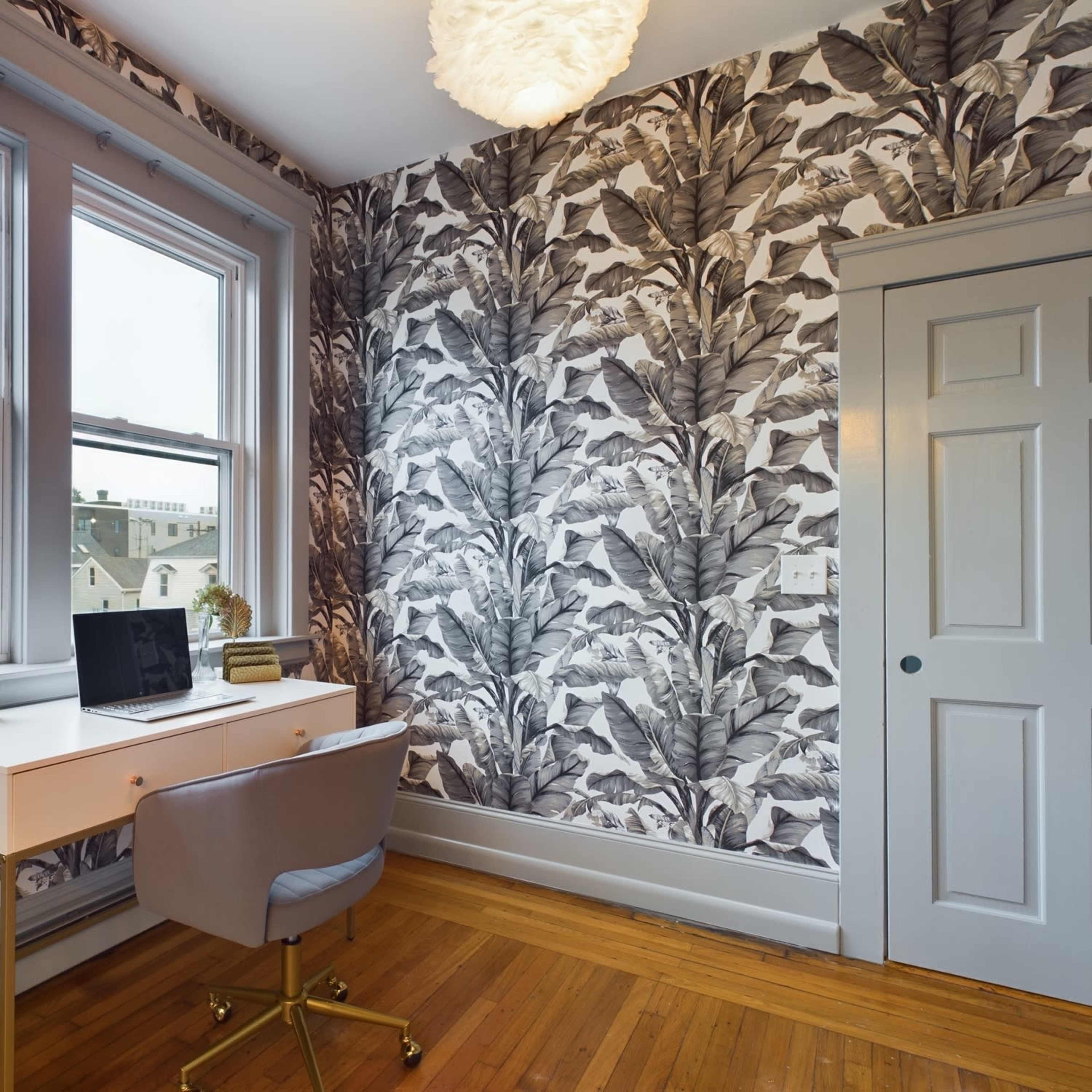 A home office features a white desk with a laptop in front of patterned wallpaper depicting large leaves, and a gray chair on a wooden floor next to a door.