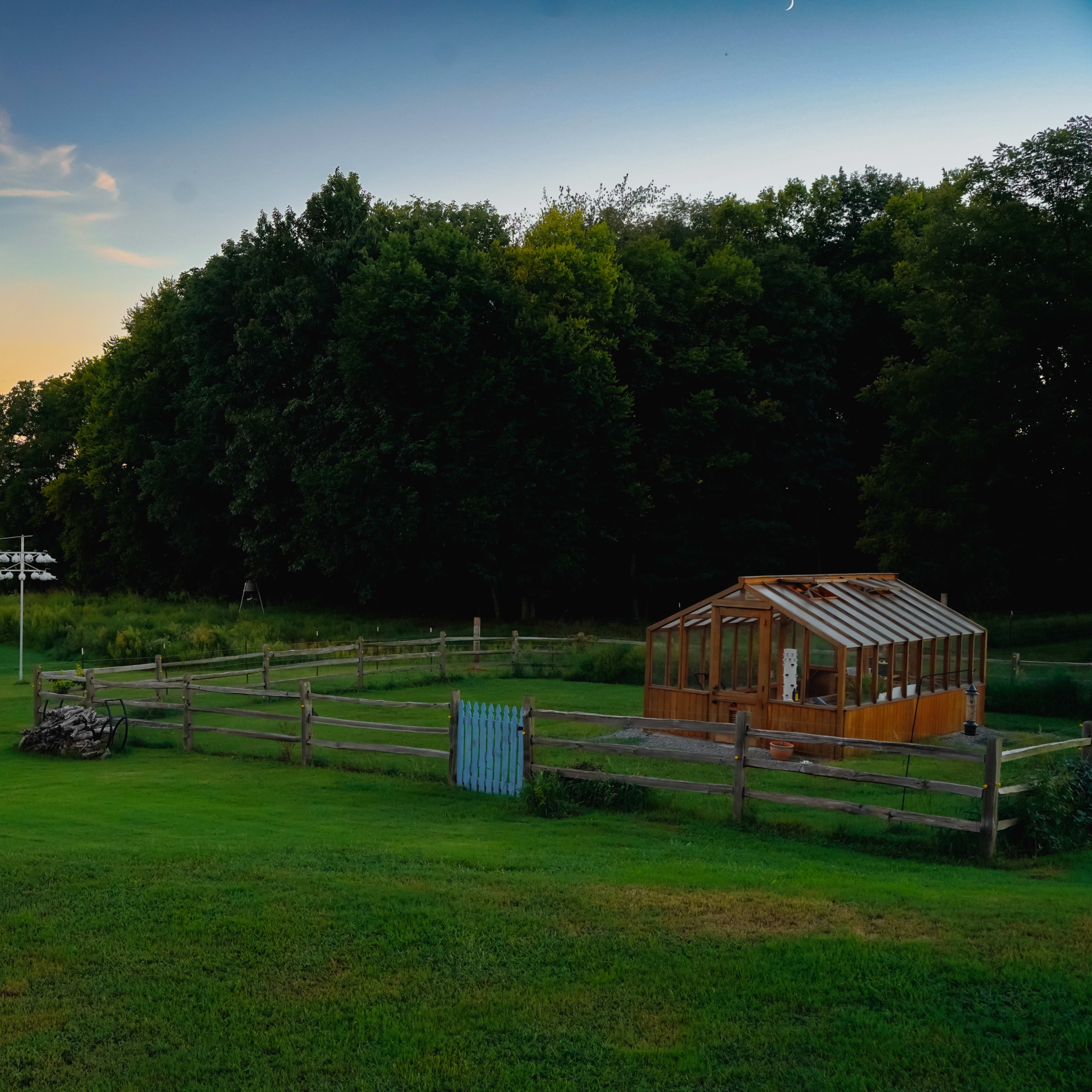 A wooden greenhouse stands beside a fenced area on a grassy landscape, with a sunset illuminating the sky and trees in the background.