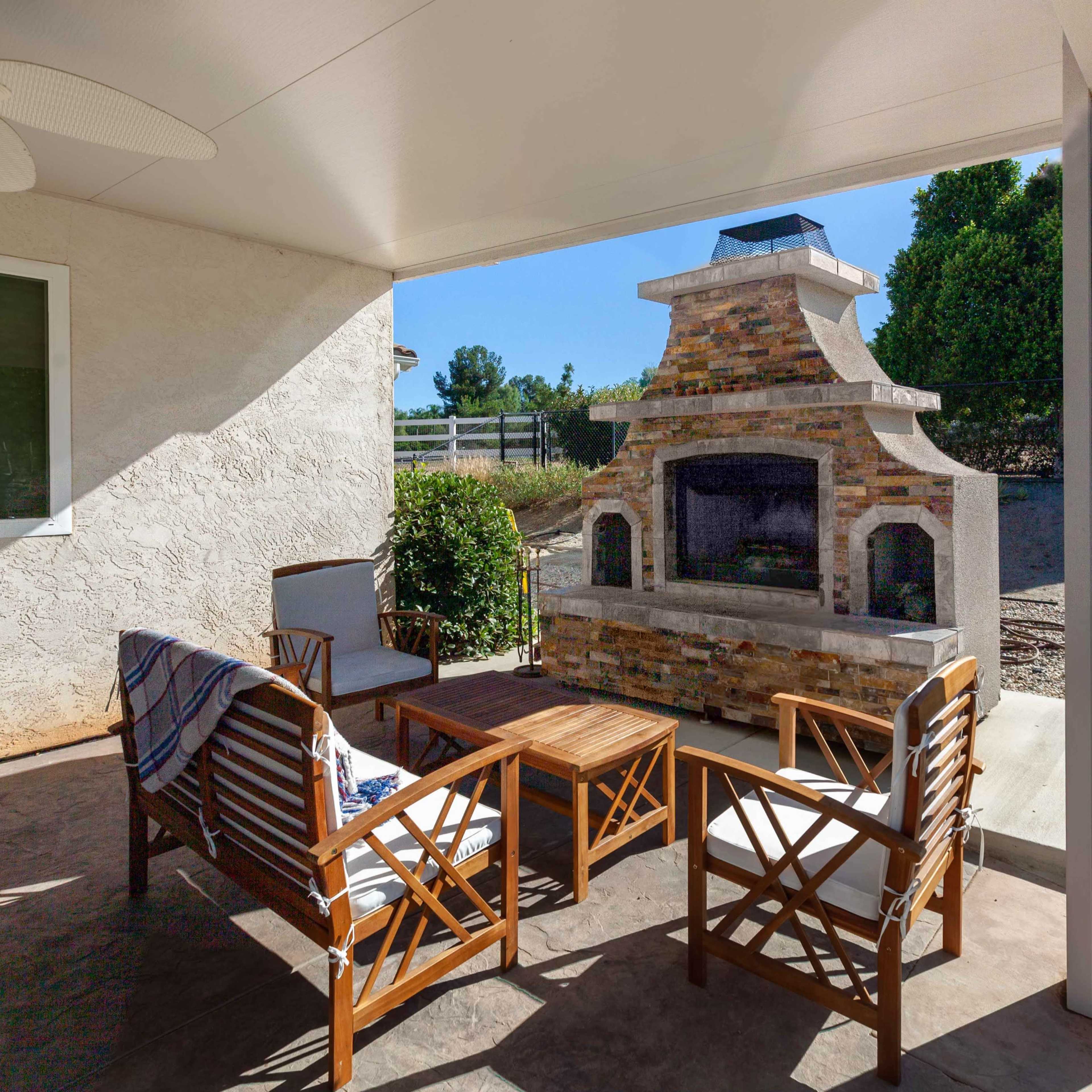 A covered patio features wooden furniture and a stone fireplace, with a grassy area and trees visible in the background.